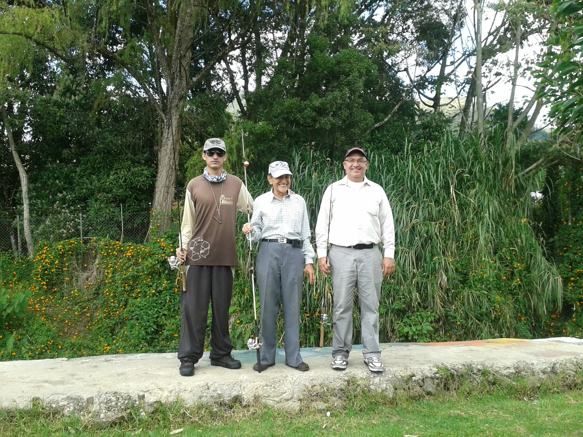 Tres generaciones de pescadores posan juntos con sus cañas en la orilla de un lago, rodeados de vegetación.