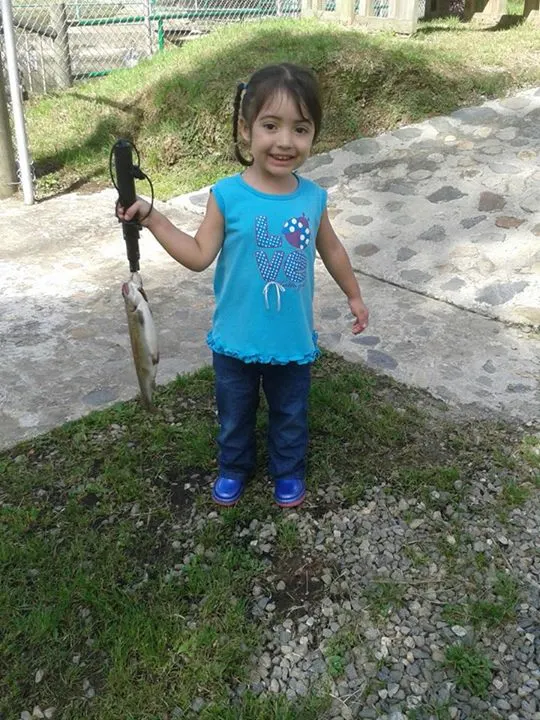Una niña pequeña con camiseta turquesa sonríe mientras sostiene una trucha pequeña colgada de una pinza.