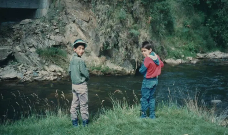 Foto antigua de dos niños pescando en la orilla de un río de montaña, junto a un peñasco cubierto de musgo.