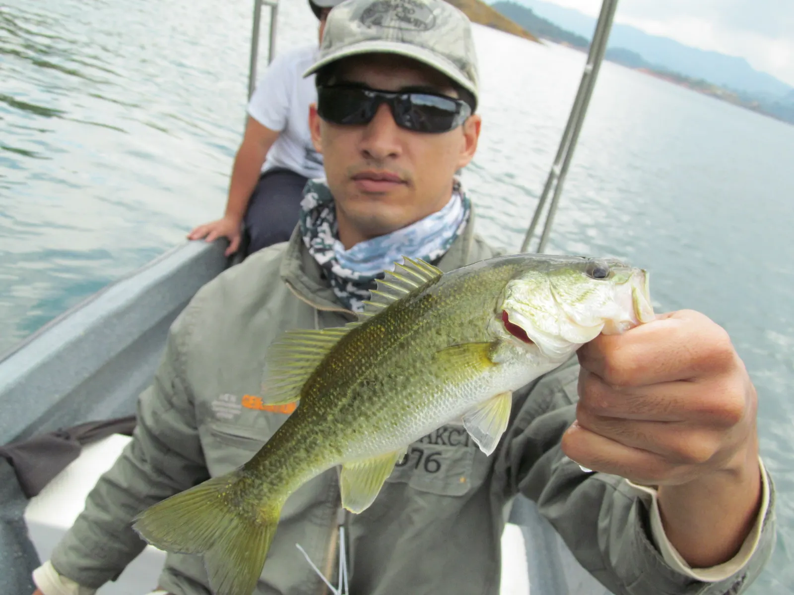 Un pescador con gorra y gafas de sol sostiene un black bass desde un bote, con el embalse gris y las montañas al fondo.