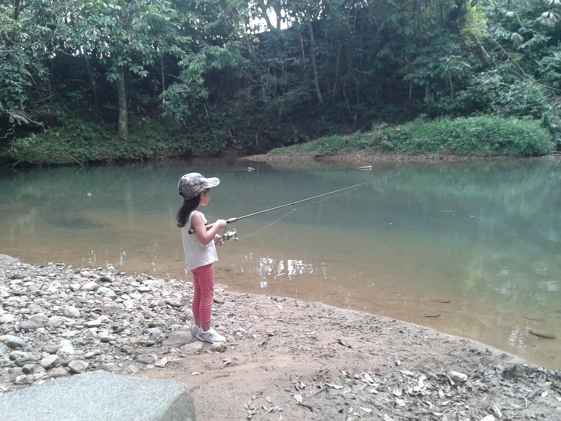 Una niña con gorra pesca de pie en la orilla pedregosa de un río de aguas claras rodeado de selva.