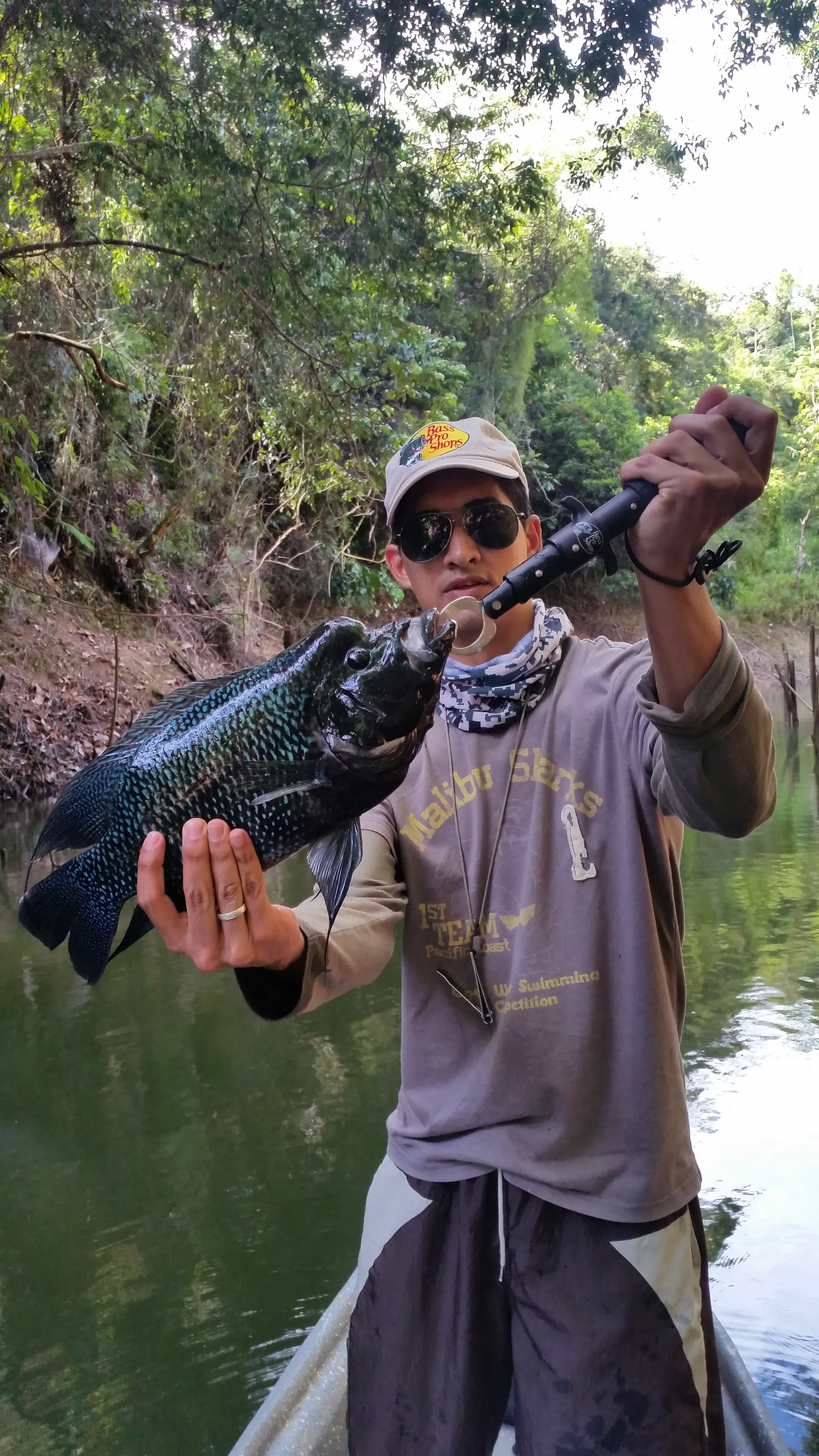 Un pescador con gorra clara y gafas de sol sostiene una mojarra negra grande de escamas azuladas, desde un bote en un brazo de río rodeado de selva.