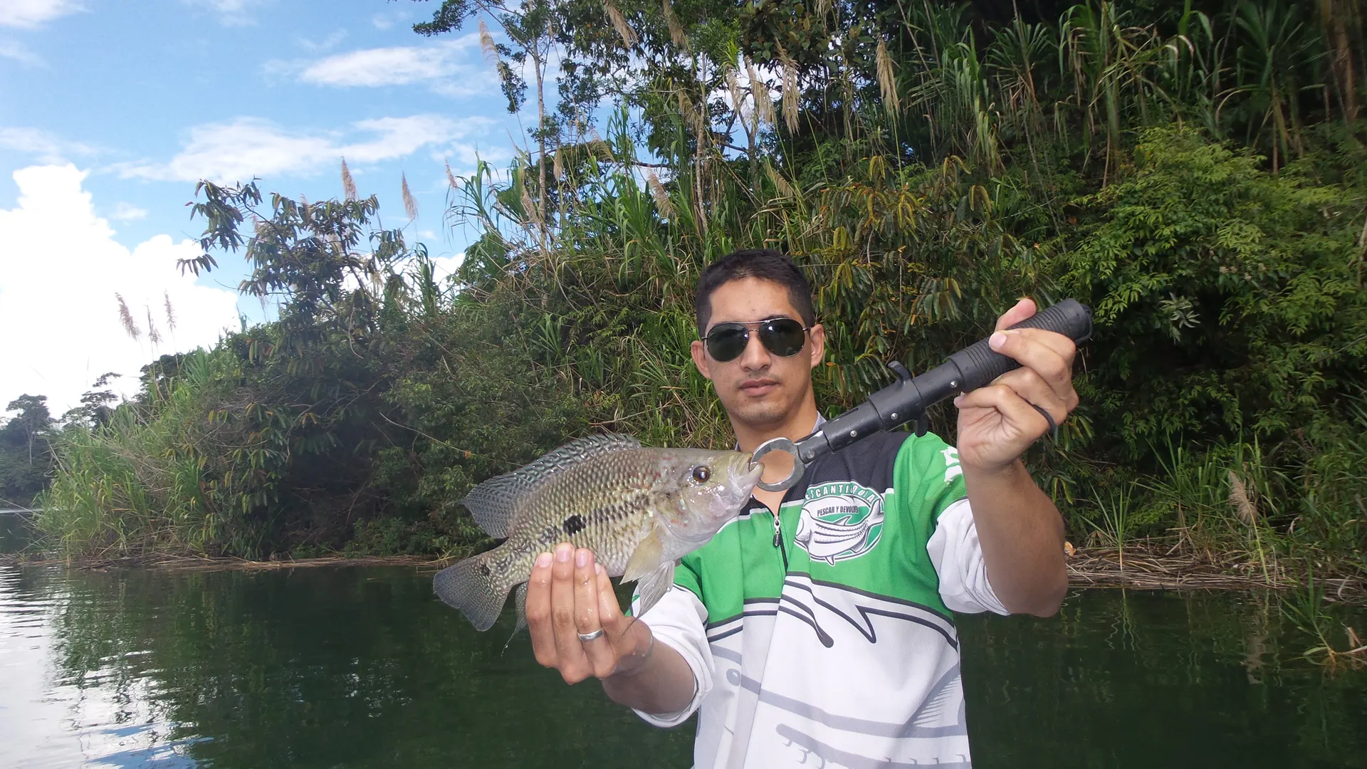 Un pescador con camiseta verde y blanca del club sostiene una mojarra sujeta con una pinza, en un brazo del embalse bordeado de vegetación.