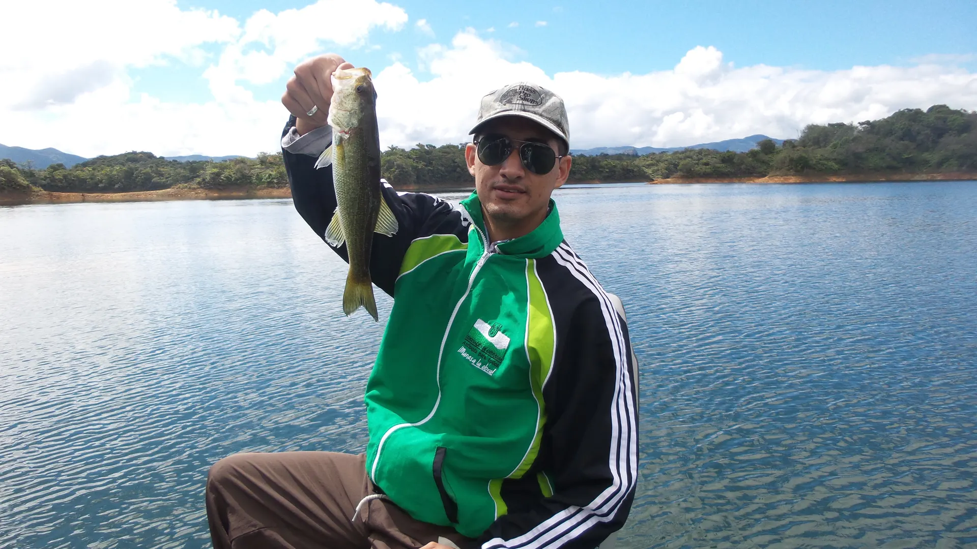 Un pescador con chaqueta verde y gafas de sol levanta un black bass pequeño desde un bote, en un embalse azul con montañas al fondo.