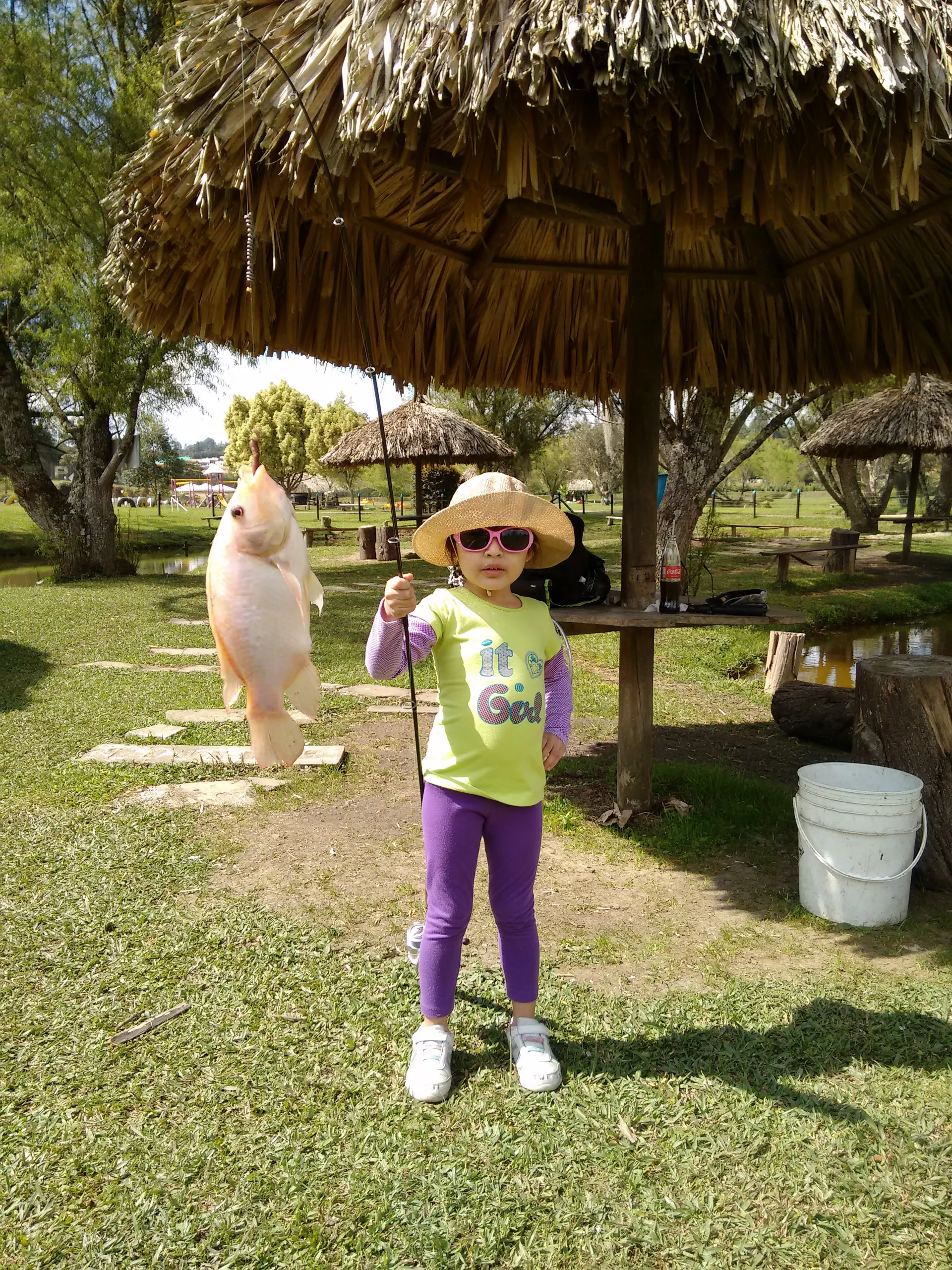 Una niña con sombrero de paja y gafas de sol rosadas sostiene una caña de la que cuelga una cachama, junto a un kiosco de palma en un lago de pesca.