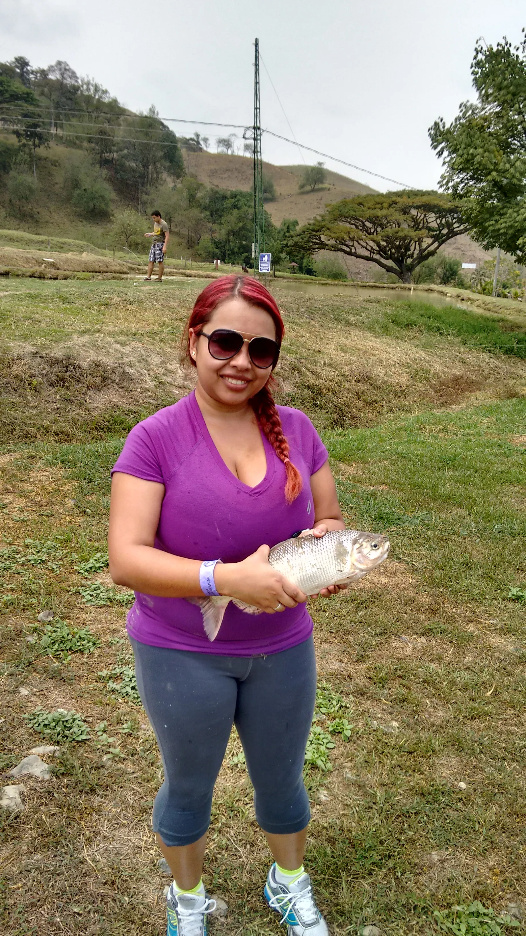Una mujer de pelo rojo con gafas de sol sonríe sosteniendo una cachama plateada, en un lago de pesca rodeado de potreros y montañas.