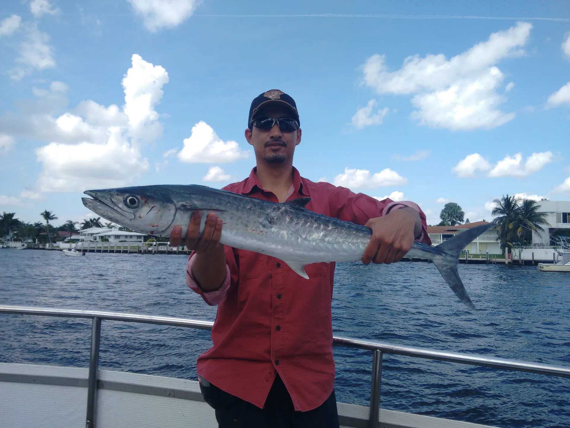 Un pescador con camisa roja sostiene una sierra plateada con las dos manos en la cubierta de un barco, con casas y palmeras al fondo.