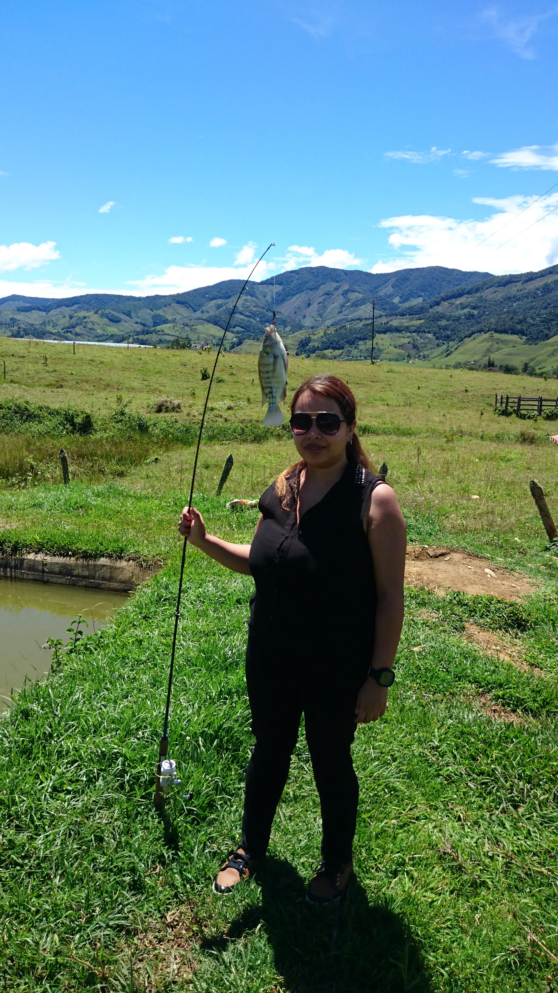 Una mujer de pie en un potrero levanta una caña de la que cuelga una mojarra, junto a un estanque, con montañas verdes al fondo.