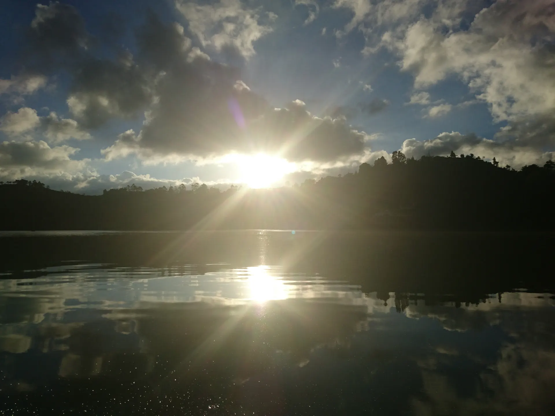 El sol poniéndose entre las nubes sobre un embalse en calma, con las montañas en silueta y todo el cielo reflejado en el agua.