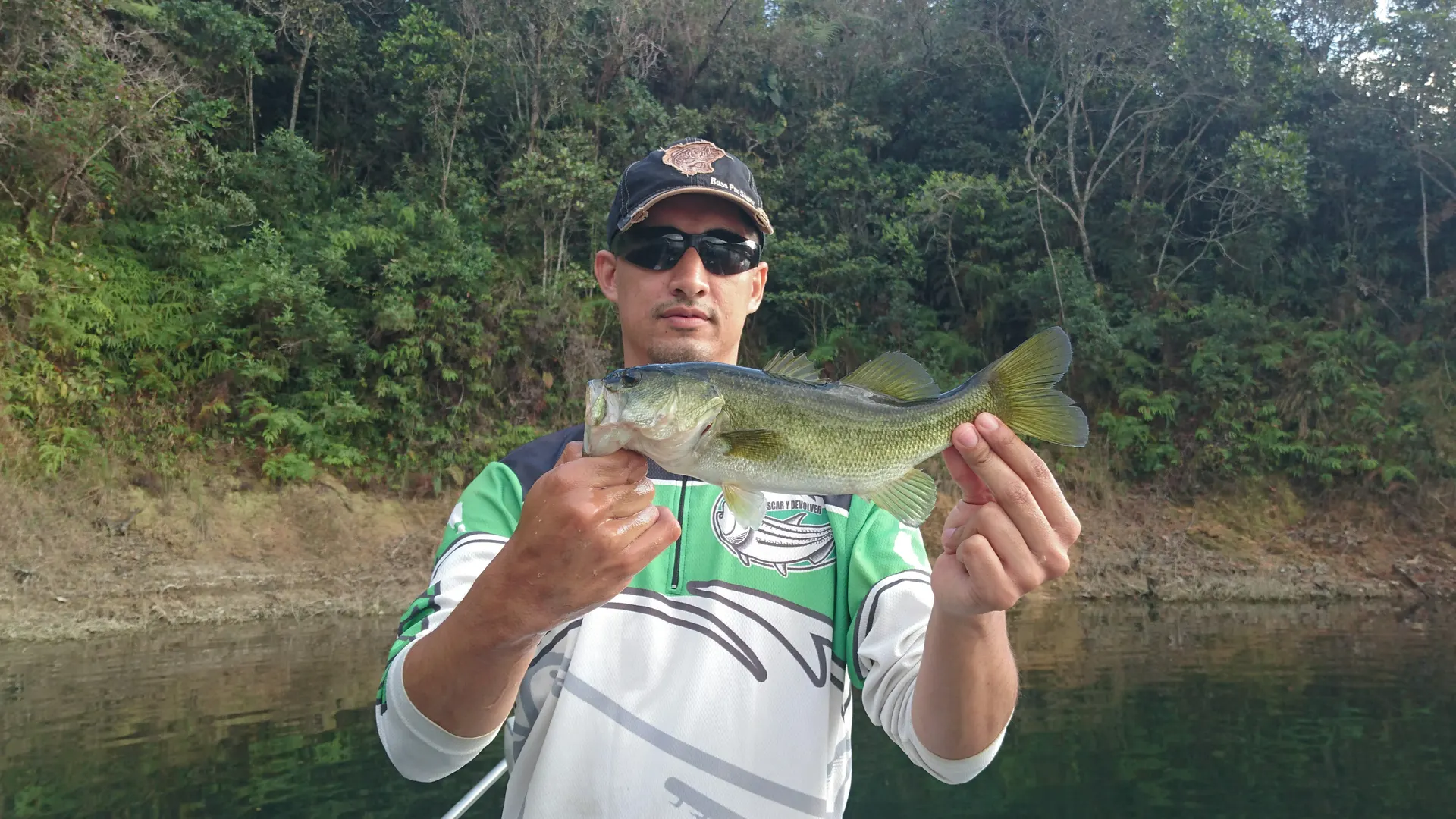 Un black bass es sostenido con una mano sobre la superficie del agua.