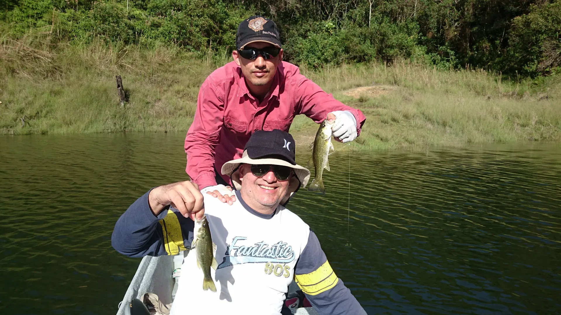 Dos pescadores en un bote sonríen mostrando cada uno un black bass pequeño, con la orilla de pasto y monte al fondo.