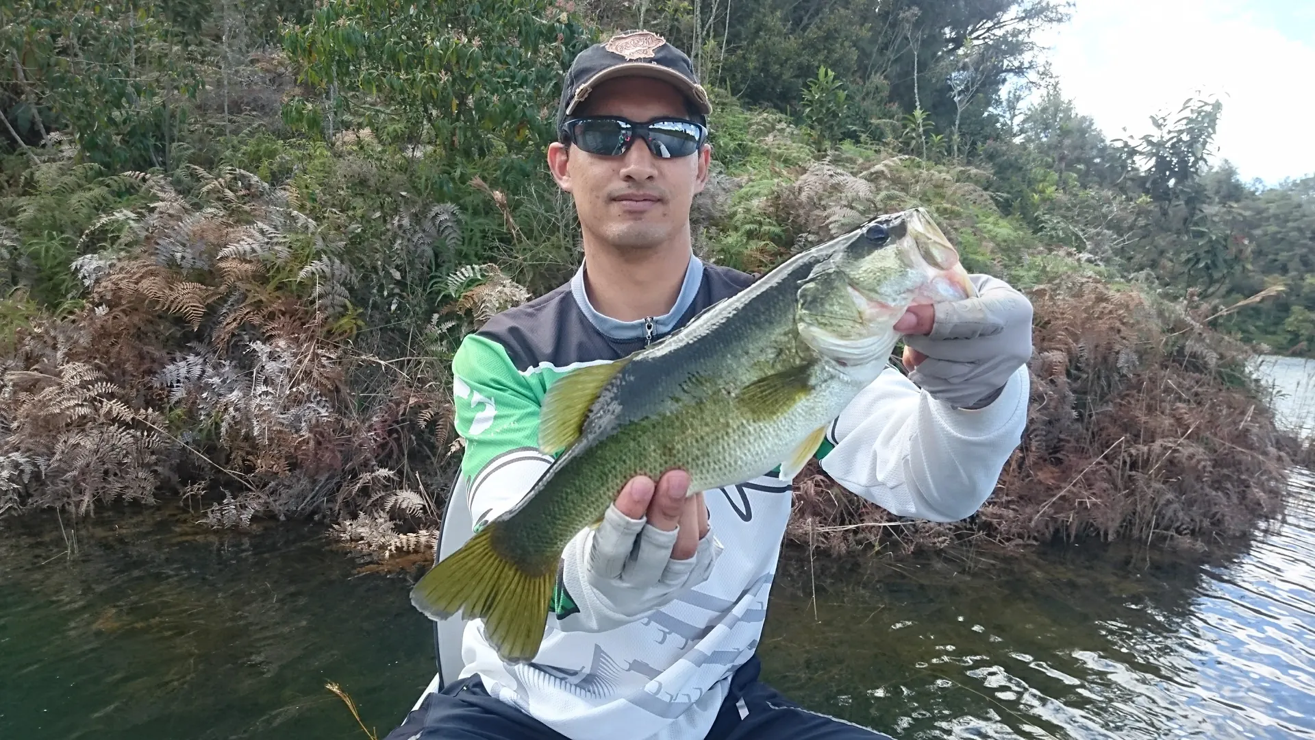 Un pescador con gorra y gafas de sol sostiene un black bass con las dos manos, junto a una orilla cubierta de helechos secos.