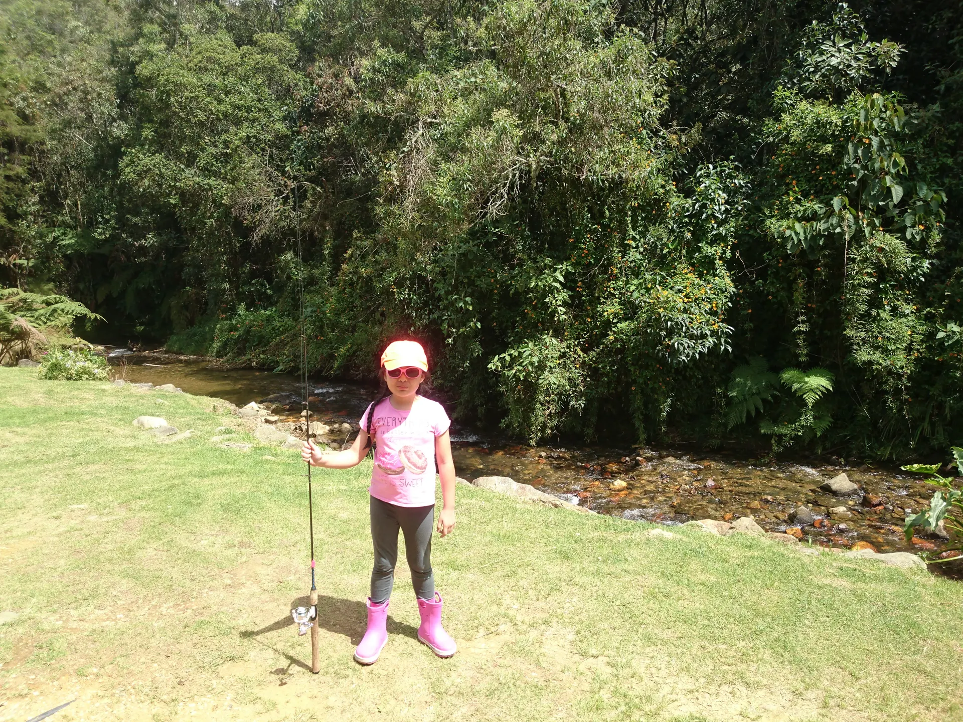 Una niña con gorra amarilla, gafas rosadas y botas de caucho posa con su caña junto a una quebrada de montaña rodeada de vegetación.