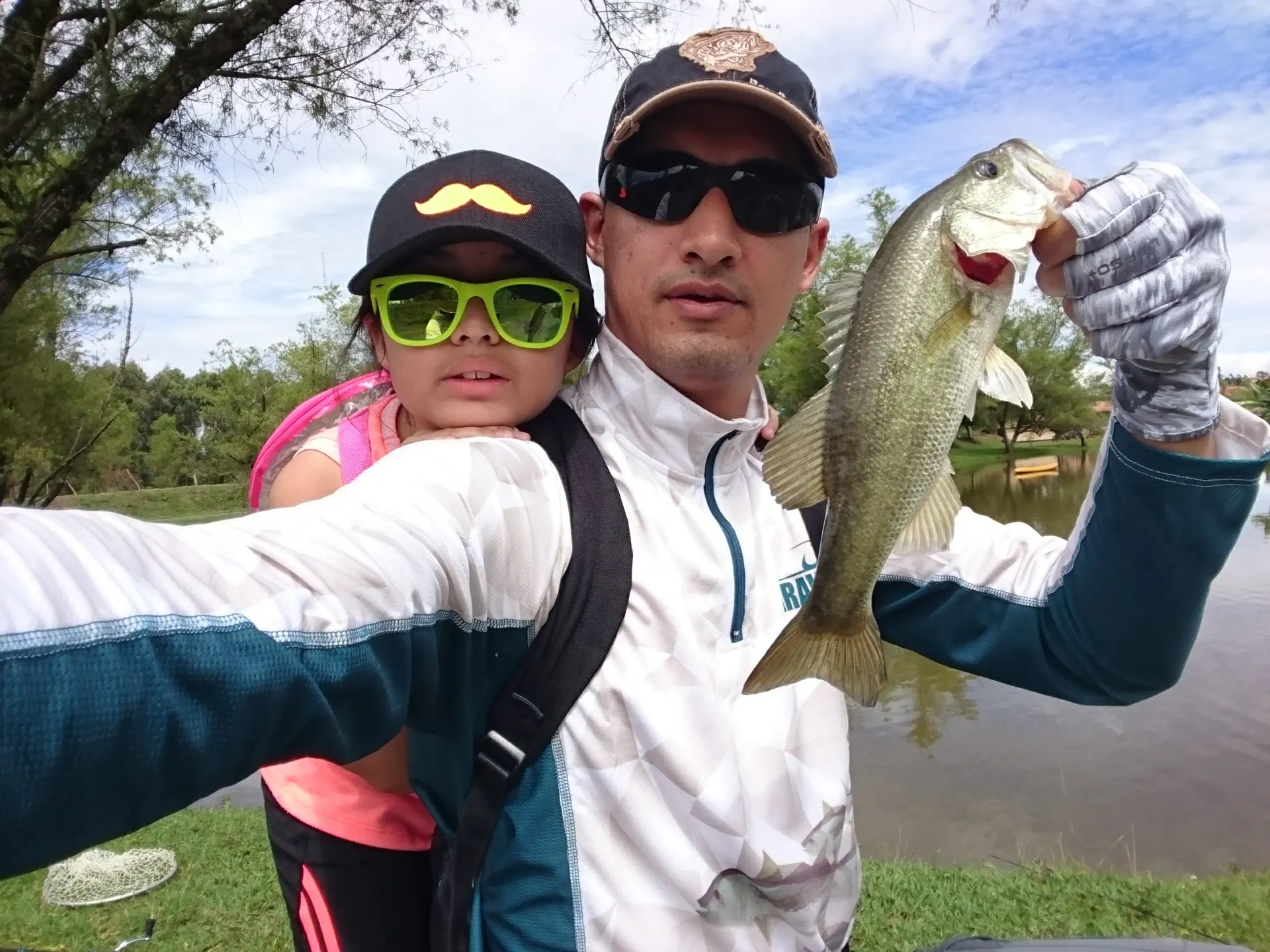 Un padre se toma una selfie con su hija cargada en la espalda mientras sostiene un black bass, a la orilla de un lago.