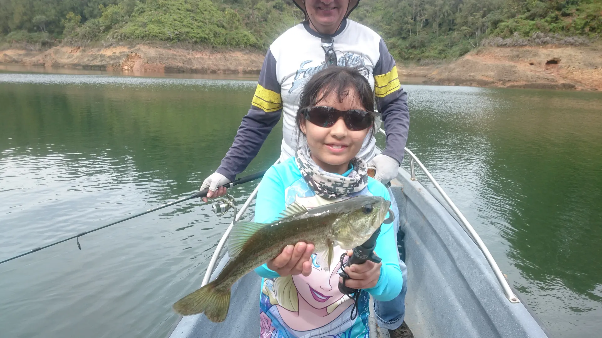 Una niña con gafas de sol sonríe sosteniendo un black bass desde un bote, mientras su abuelo pesca detrás de ella en el embalse.