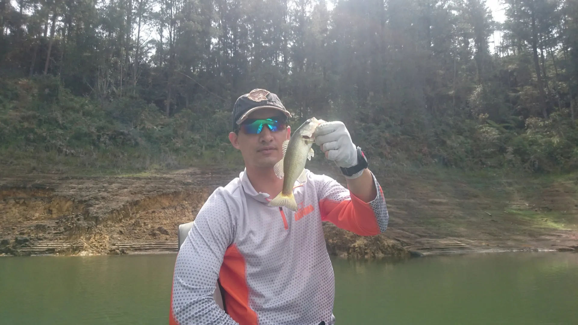 Un pescador con gorra y gafas de espejo levanta un black bass pequeño con guante blanco, con la orilla boscosa del embalse detrás.