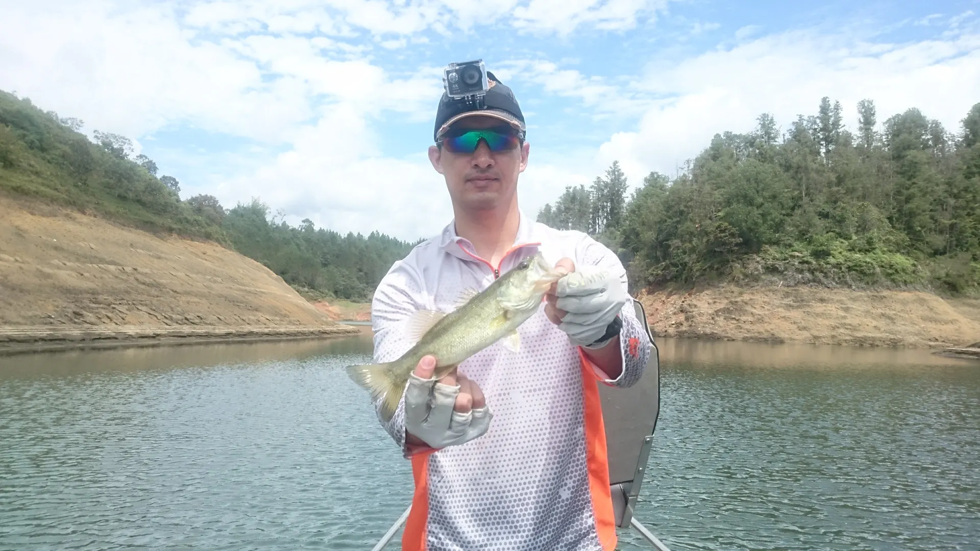 Un pescador con cámara sujeta a la gorra sostiene un black bass con las dos manos, en la proa de un bote rodeado de pinos.
