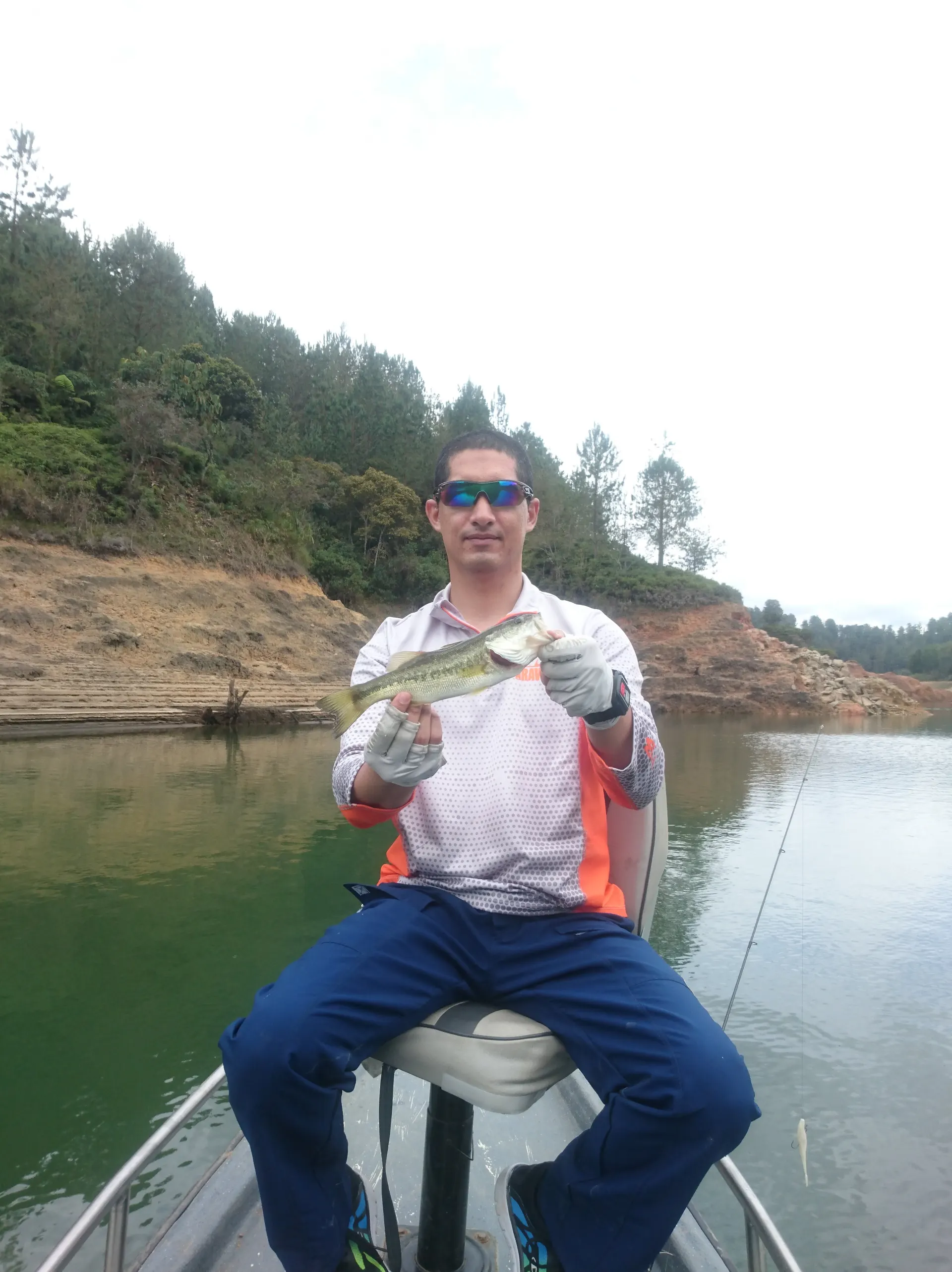 Un pescador con gafas de espejo sostiene un black bass pequeño sentado en la silla del bote, con laderas de tierra roja y pinos al fondo del embalse.