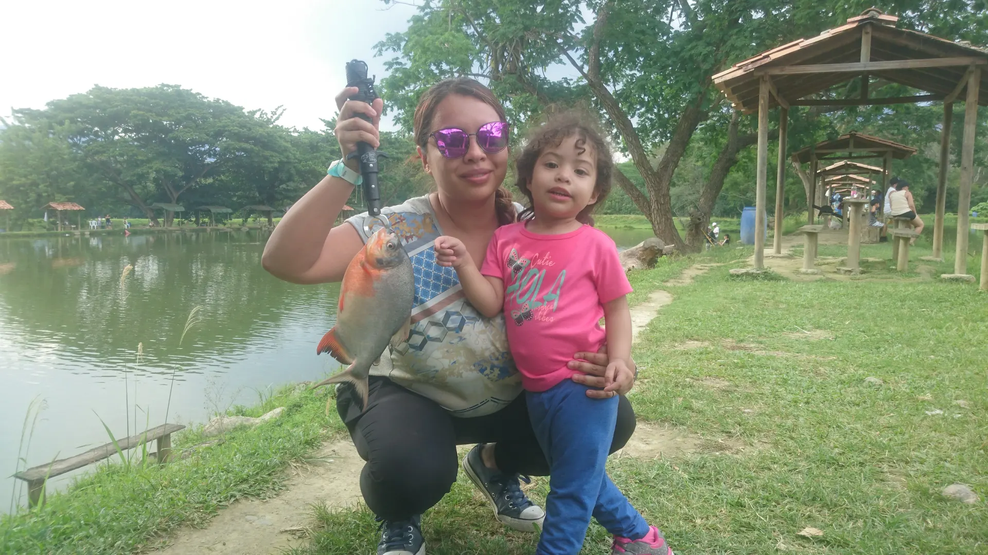 Una mujer agachada junto a su hija pequeña muestra una cachama de aletas anaranjadas, a la orilla de un lago de pesca con kioscos de madera.