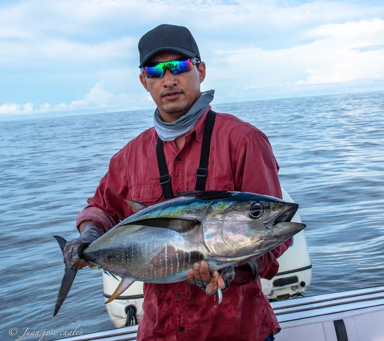 Un pescador con camisa roja, gorra y gafas de sol sostiene un atún en la cubierta de una lancha, con el mar abierto detrás.