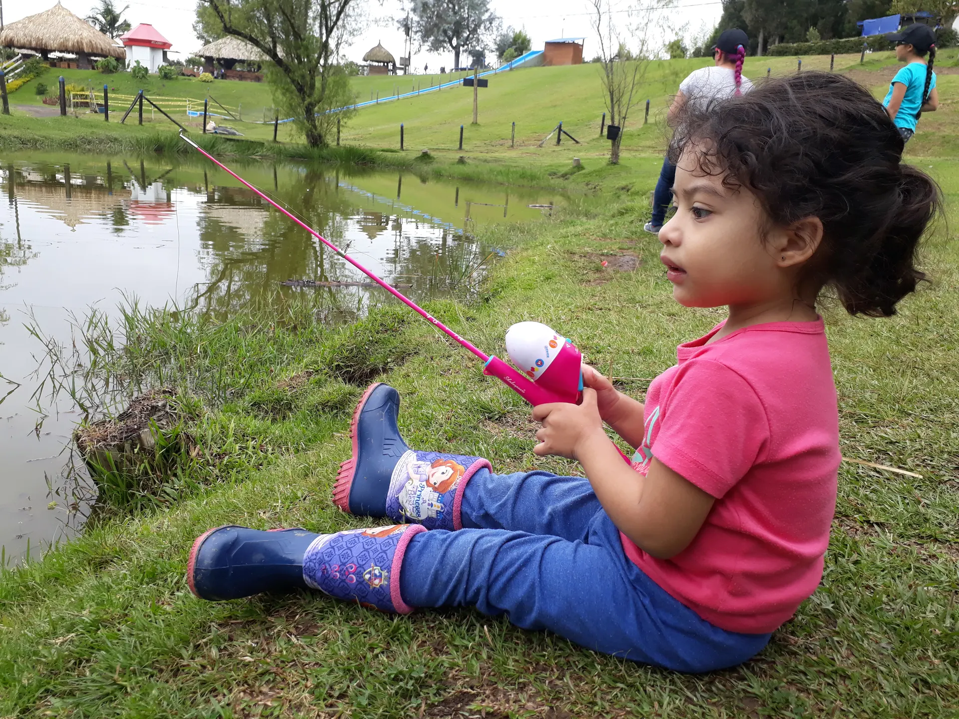 Una niña pequeña sentada en el pasto a la orilla de un lago sostiene una caña de pescar rosada, concentrada en el agua.