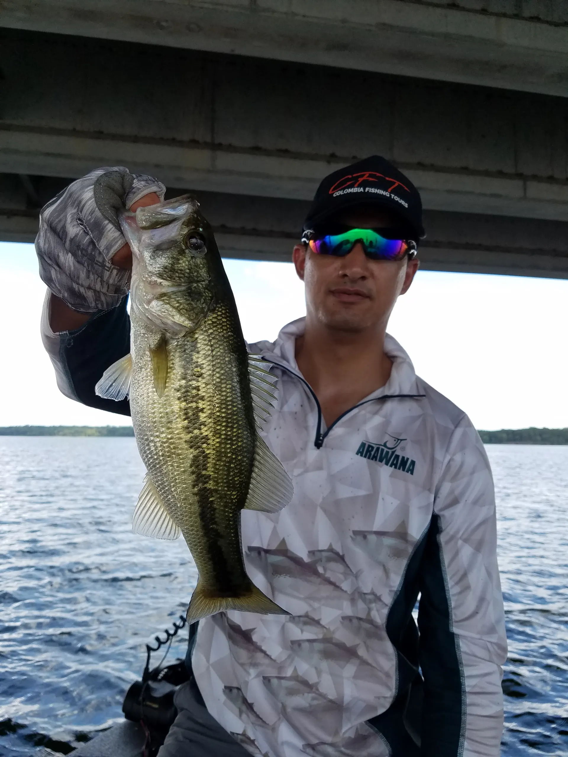 Un pescador con gorra y gafas de sol levanta un black bass bajo un puente de concreto, sobre un lago amplio.