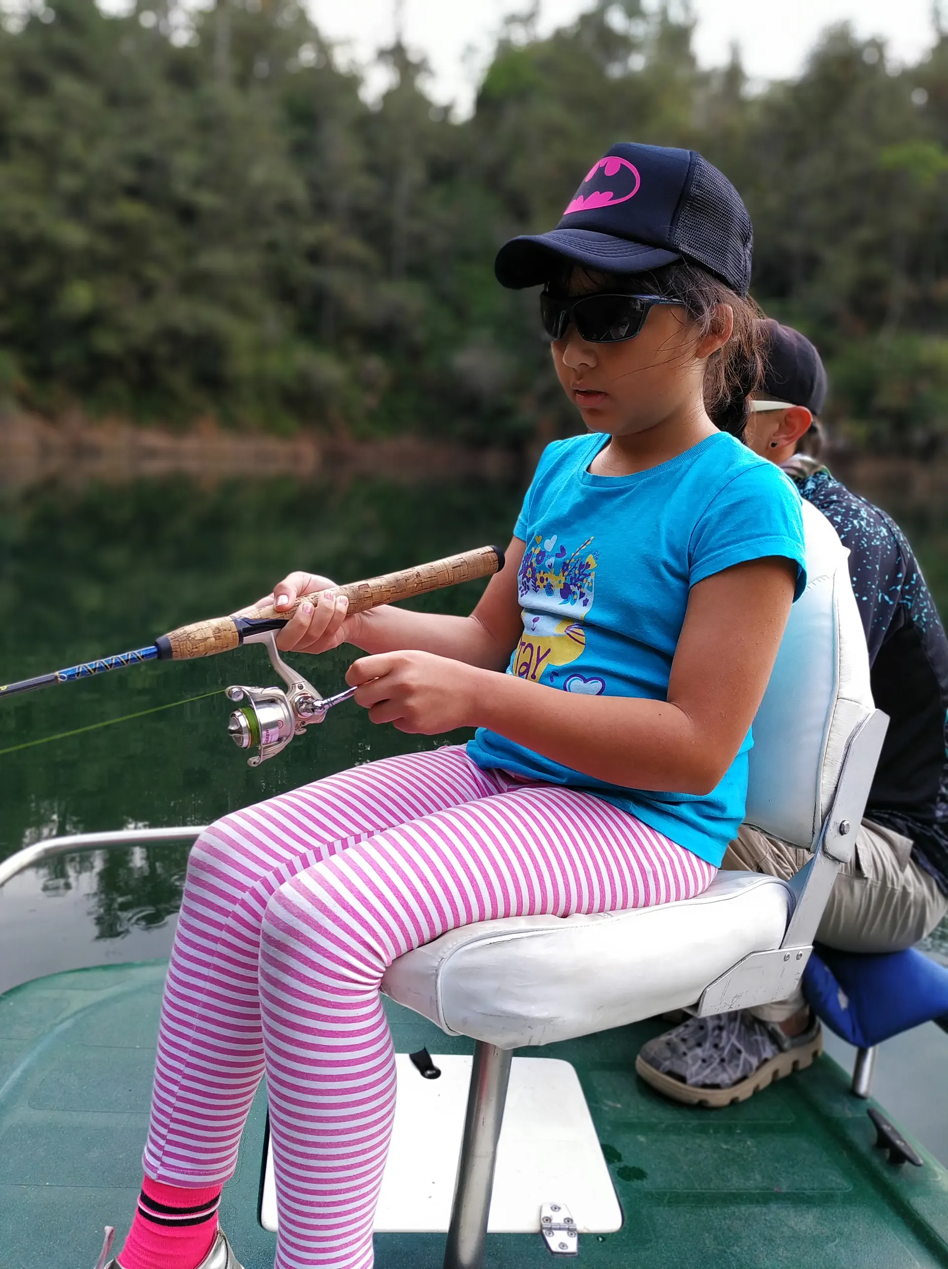 Una niña con gorra de Batman y gafas de sol recoge concentrada el carrete de su caña, sentada en la silla de un bote.