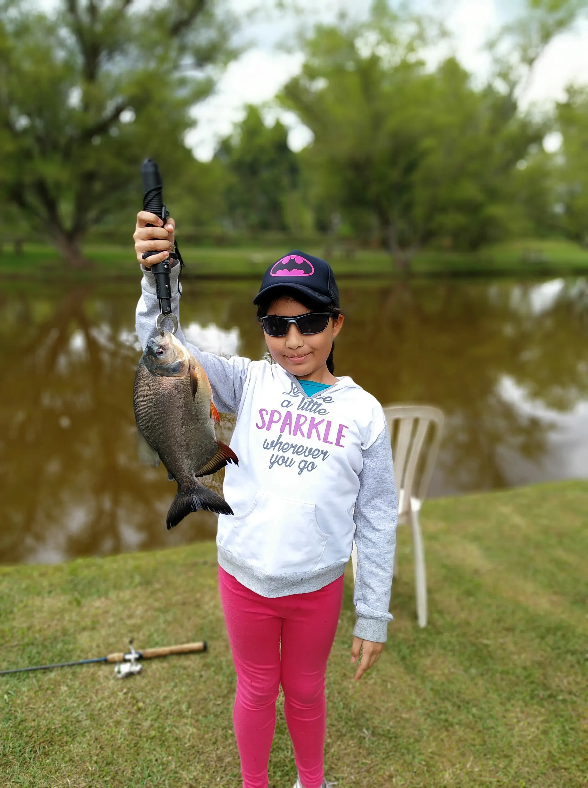 Una niña con gorra de Batman levanta con una pinza una cachama, junto a un lago de aguas color té con sillas plásticas en la orilla.