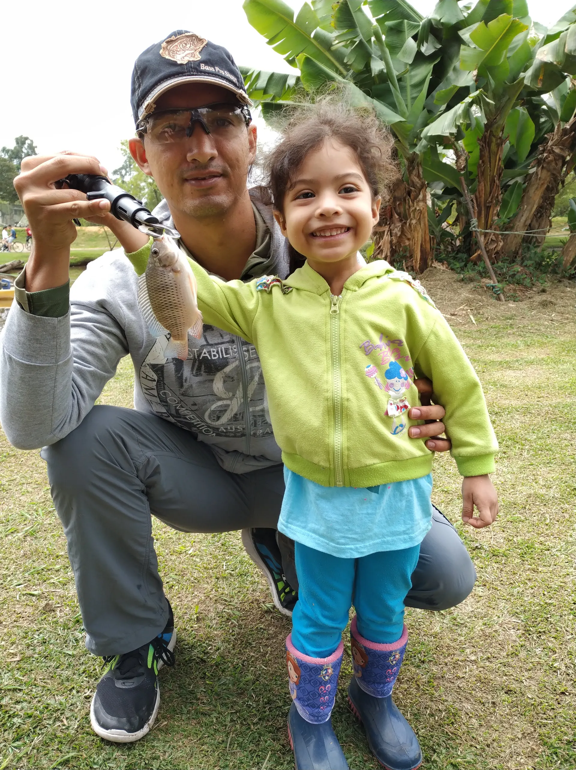 Un padre agachado junto a su hija pequeña levanta una tilapia colgada del anzuelo, frente a unas plataneras.