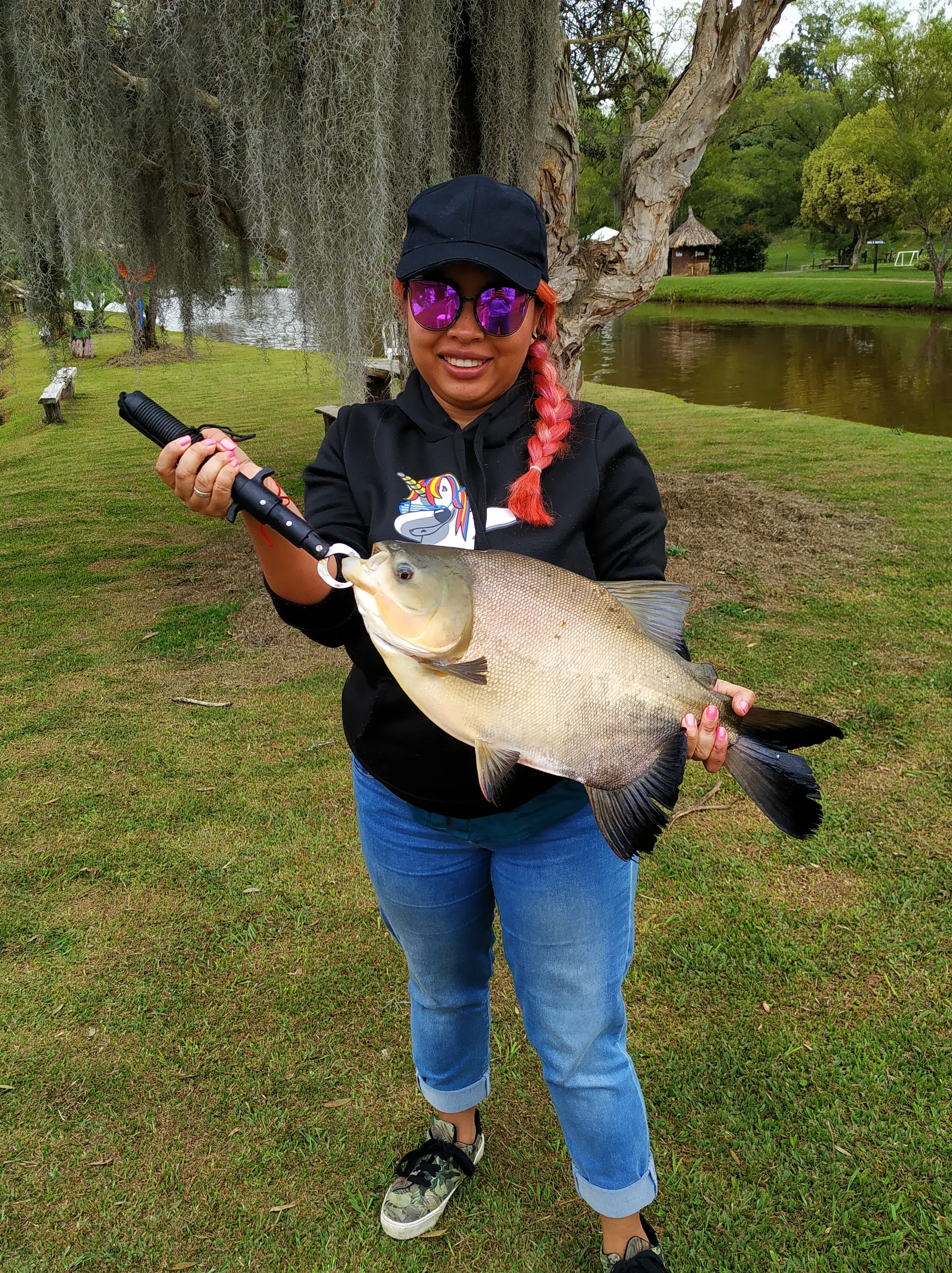 Una mujer con gorra negra, gafas de sol y trenza rosada sostiene con las dos manos una cachama grande, junto a un lago rodeado de árboles.