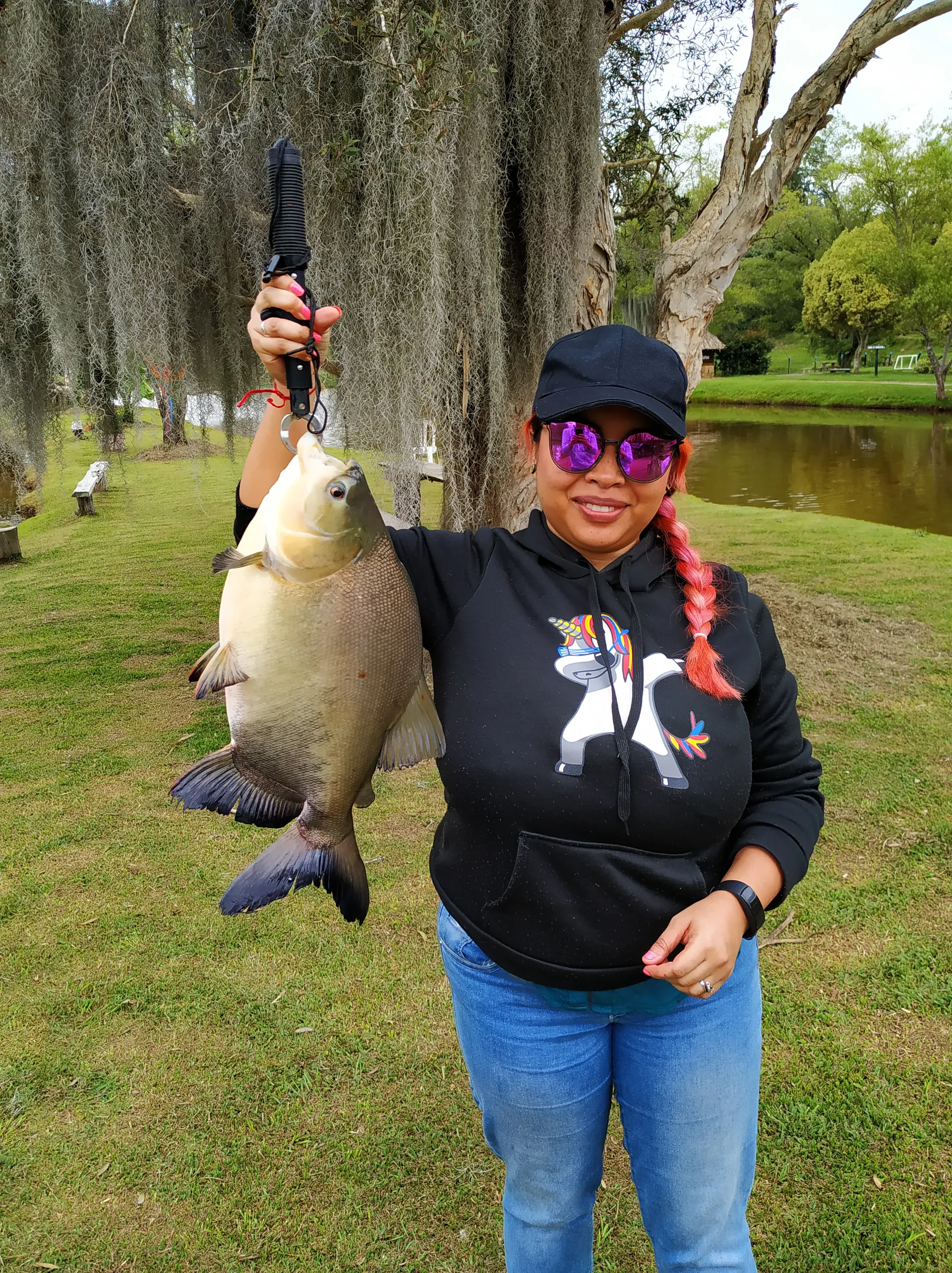 Una mujer con gorra negra, gafas de sol y trenza rosada levanta una cachama sostenida con una pinza, junto a un lago rodeado de árboles.