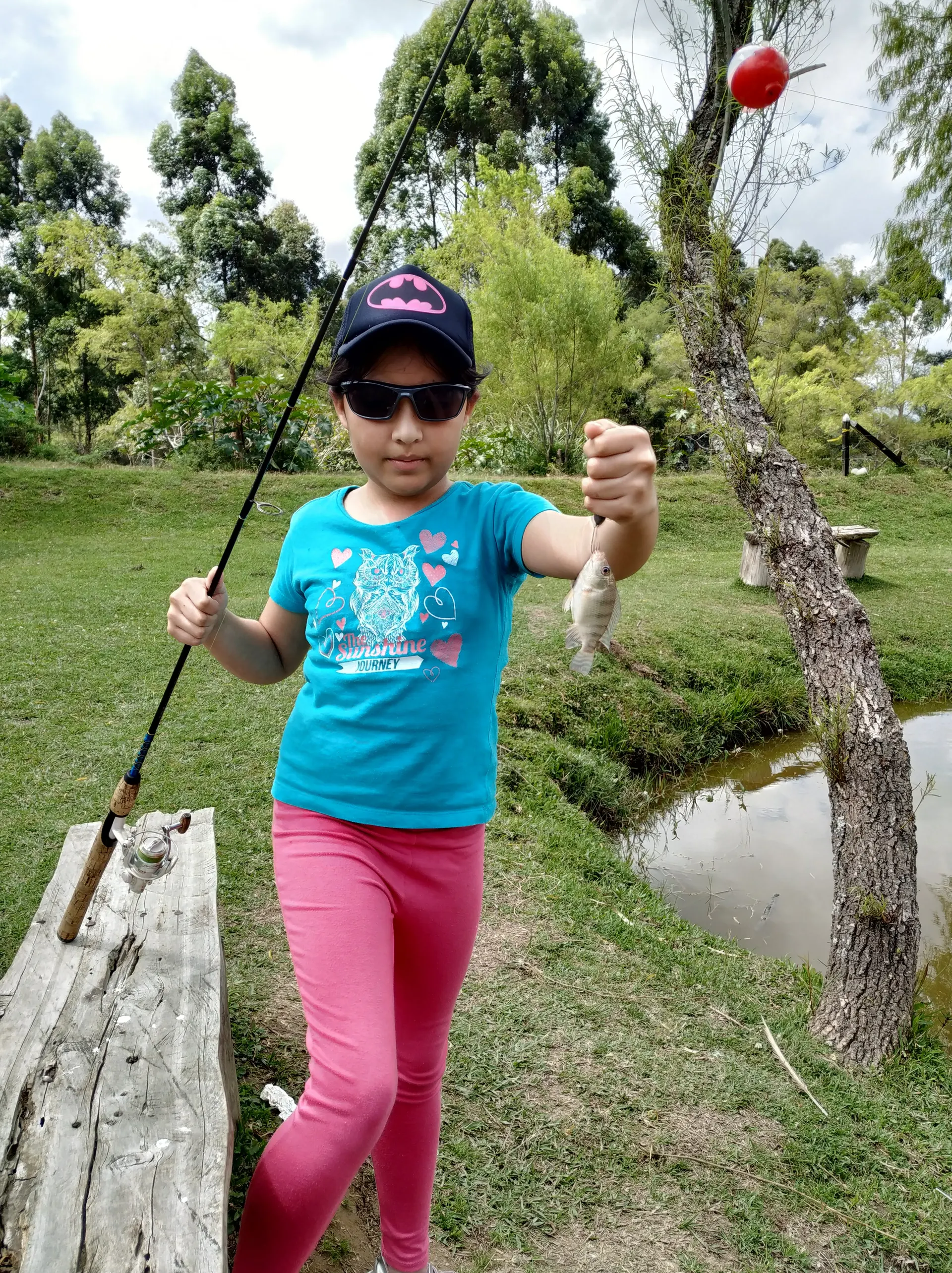 Una niña con gorra de Batman y gafas de sol levanta una tilapia pequeña colgada de la mano, junto a un estanque con boya roja.