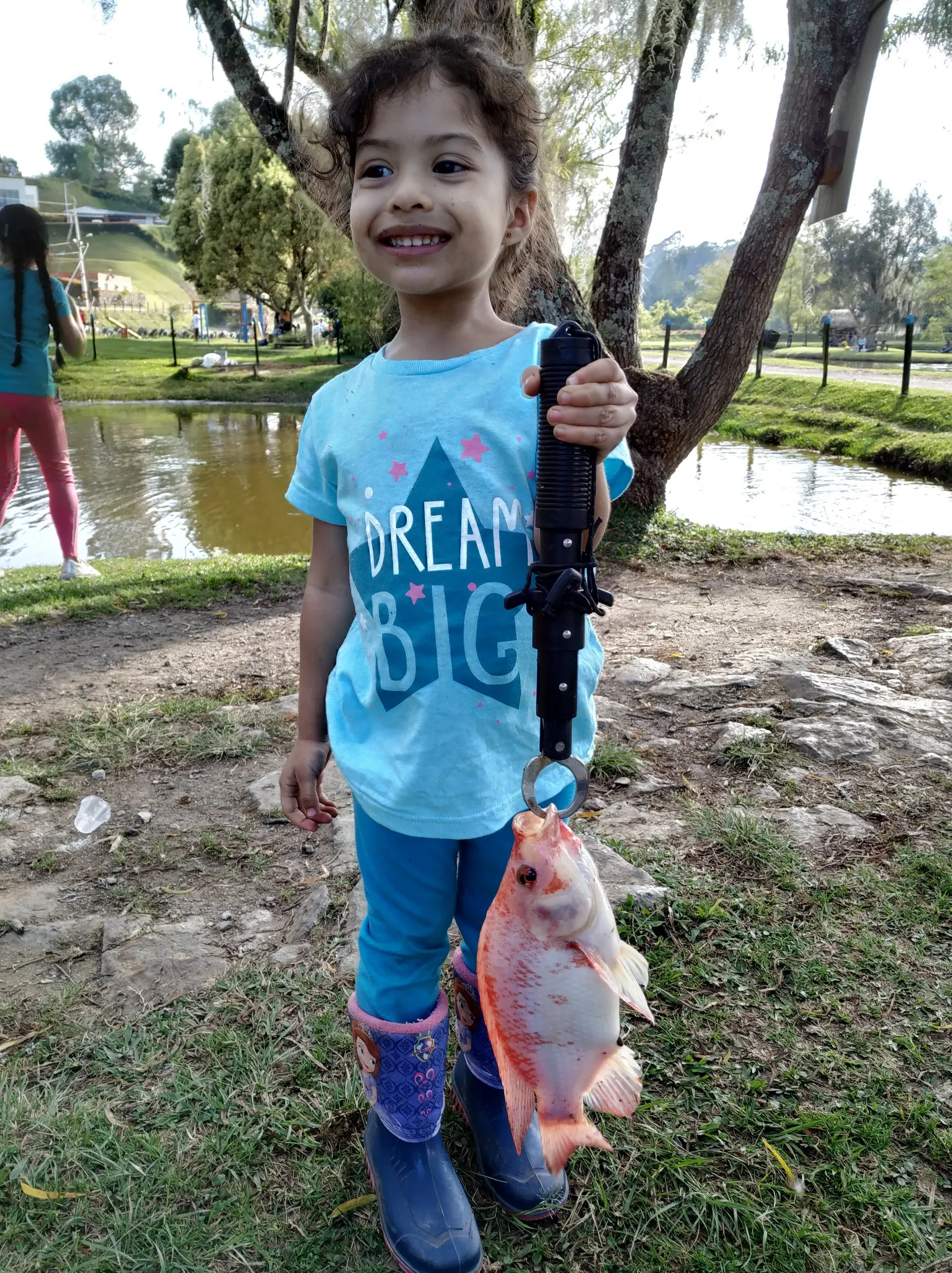 Una niña pequeña con chaqueta verde sonríe sosteniendo una pinza con una mojarra roja colgando, junto a un lago rodeado de plataneras.
