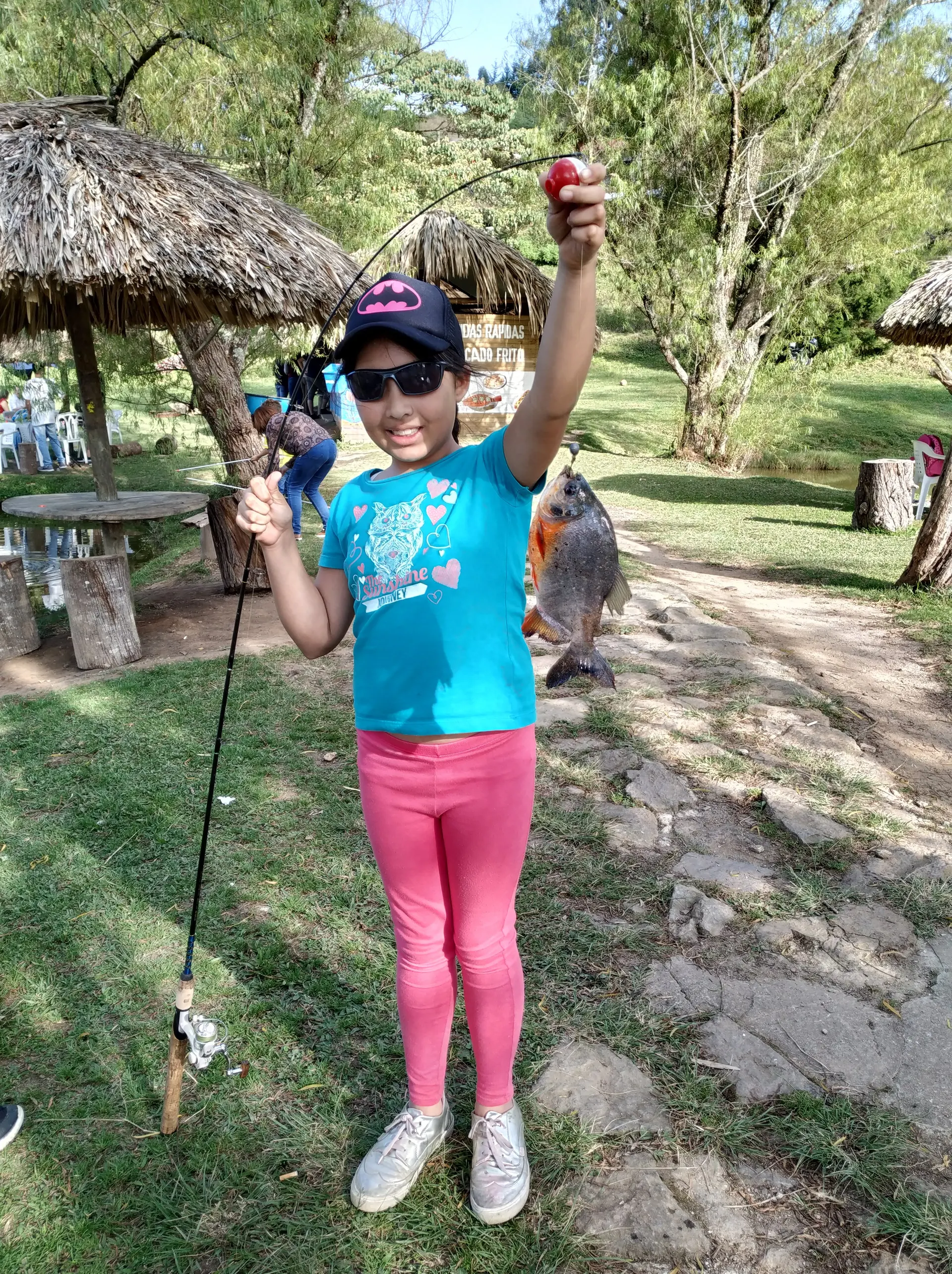 Una niña con gorra de Batman levanta el brazo mostrando una cachama plateada colgada del anzuelo, junto a kioscos de paja.