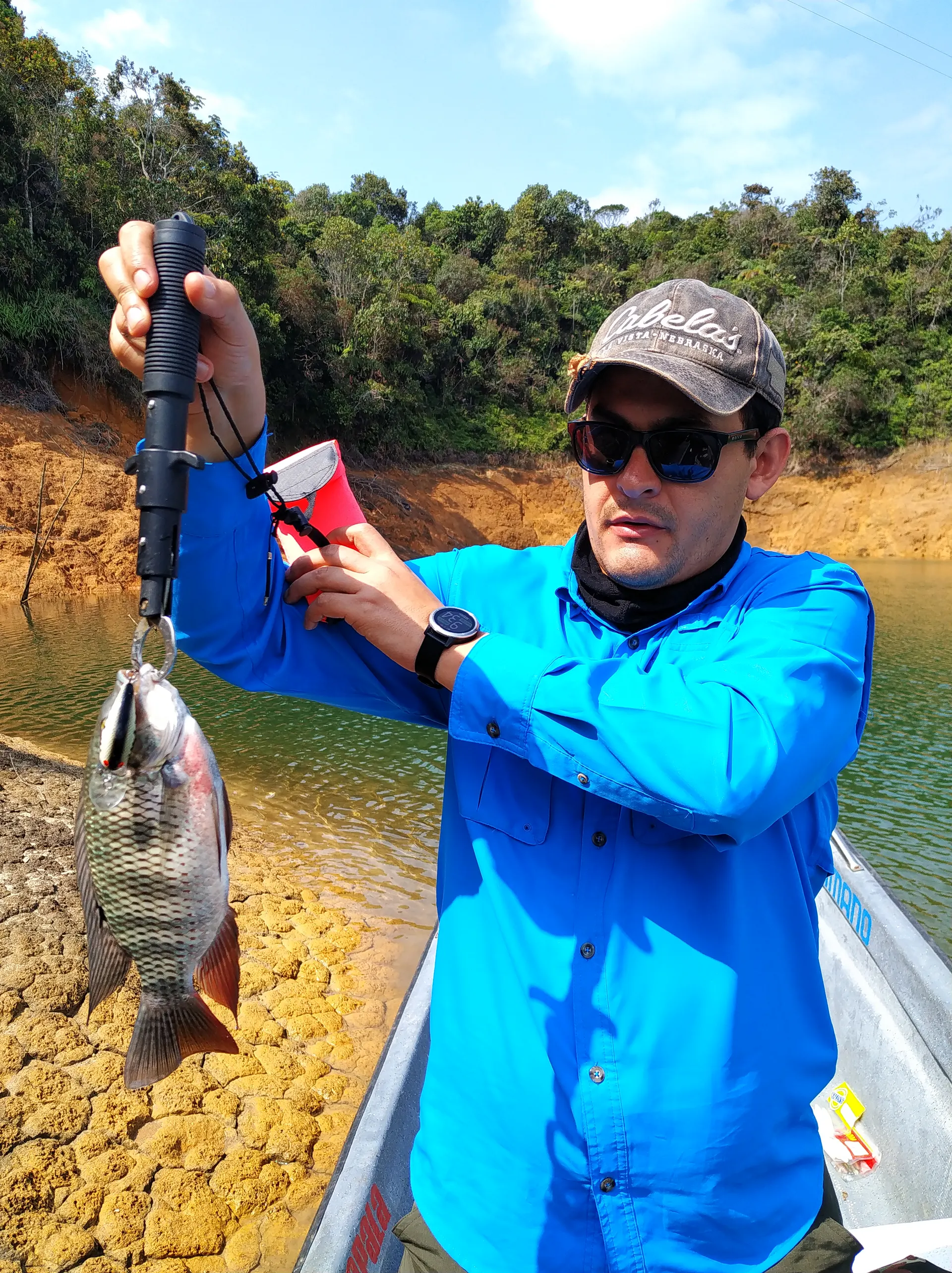 Un pescador con gorra y camisa azul levanta una tilapia colgada de un señuelo, junto a la orilla de tierra amarilla del embalse.