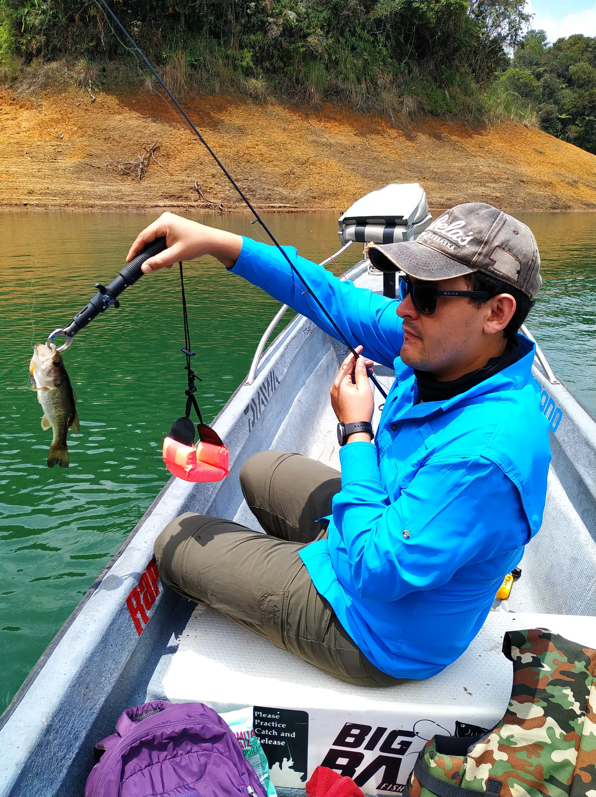 Un pescador sentado en el bote sostiene la caña con un black bass pequeño colgado del señuelo, junto a la orilla del embalse.