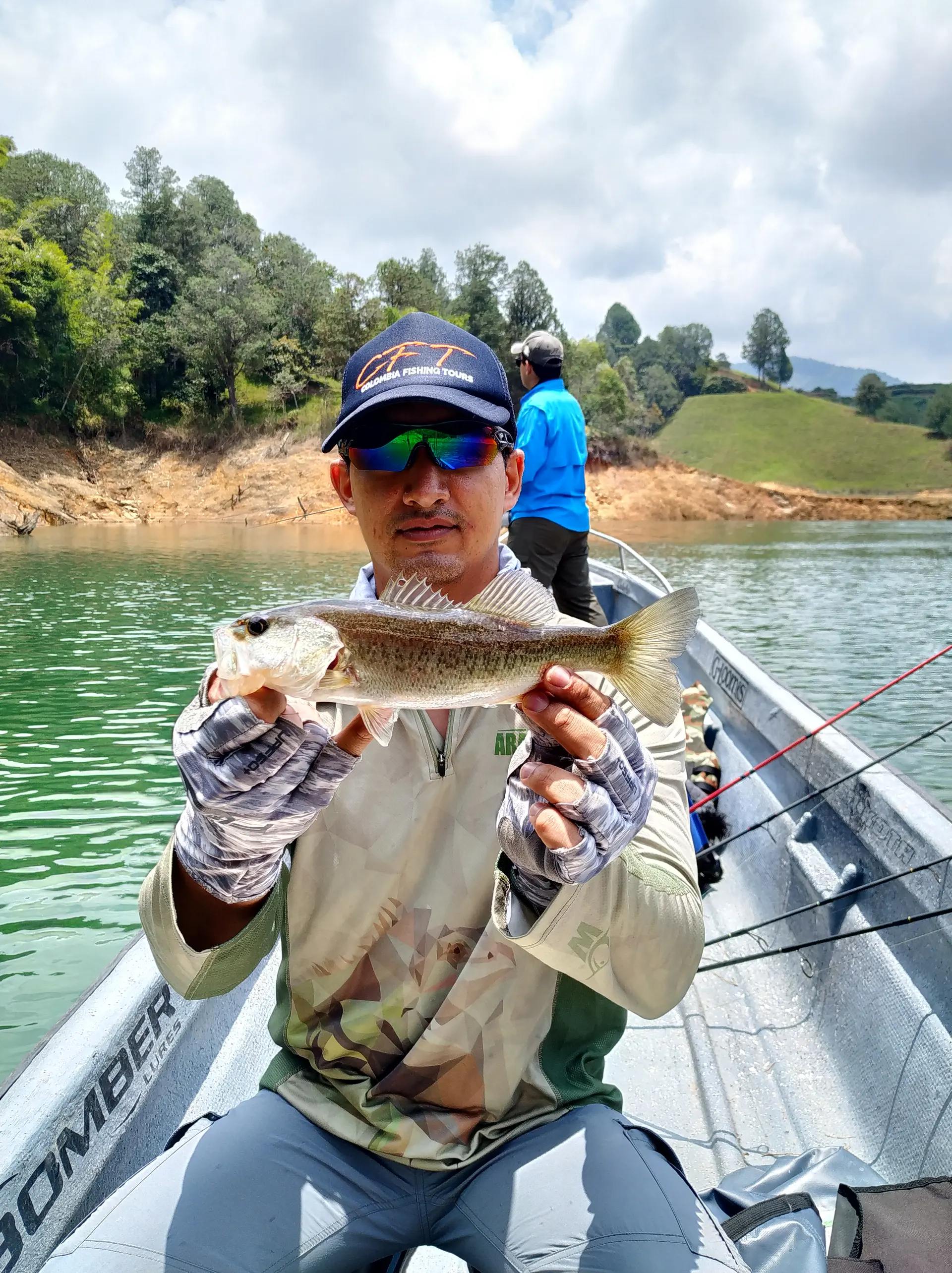 Un pescador con gafas de espejo sostiene un black bass con las dos manos, con otro pescador y las colinas del embalse detrás.