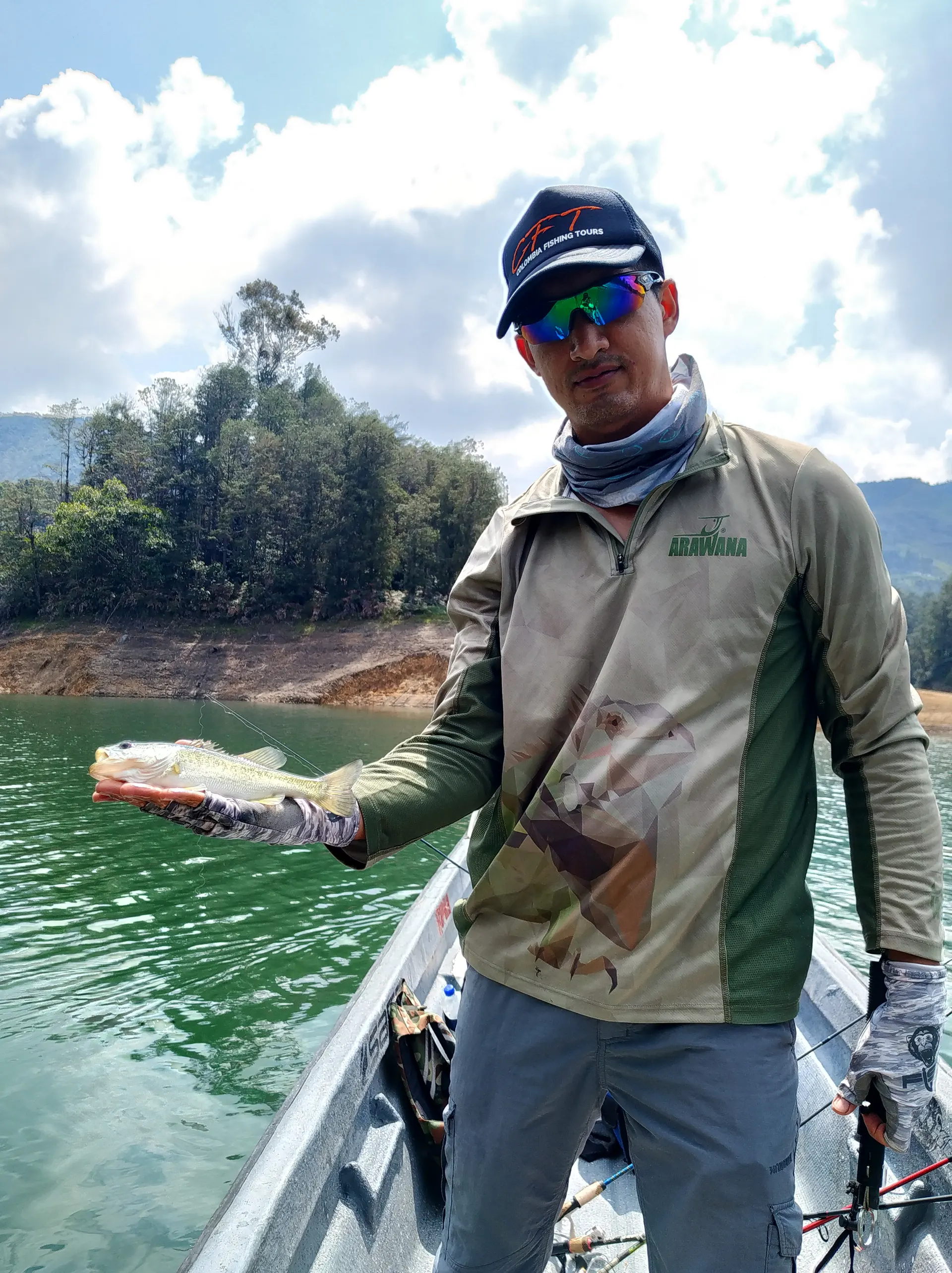 Un pescador con gorra oscura sostiene un black bass pequeño con una mano extendida, con otro pescador y colinas verdes al fondo.