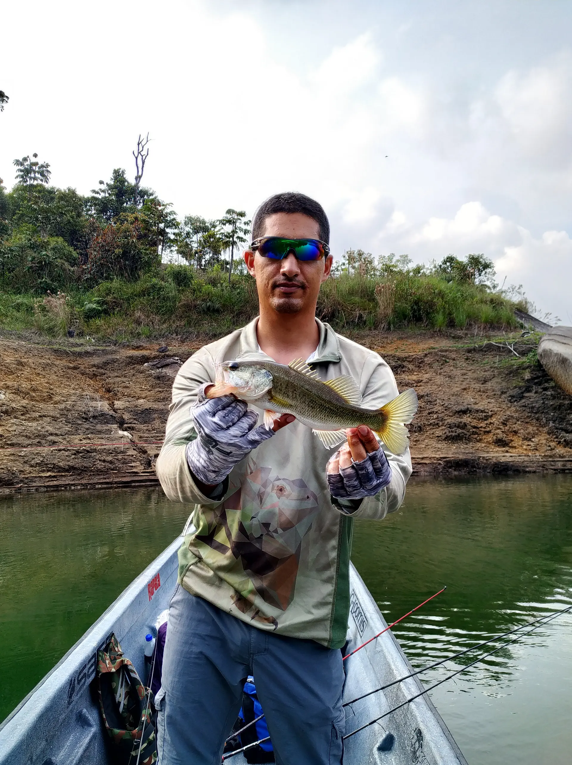 Un pescador con gafas de espejo sostiene con las dos manos un black bass, en la proa de un bote sobre el embalse con orillas de tierra roja.