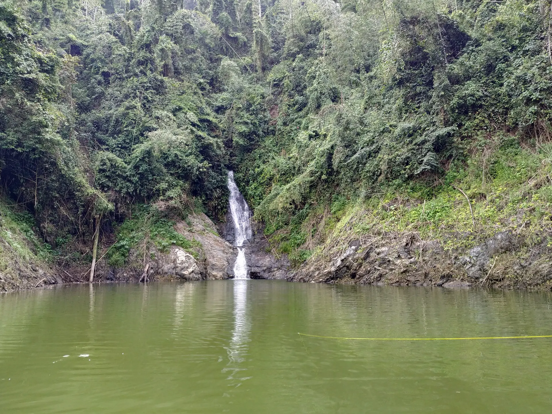 Una cascada angosta cae entre rocas y vegetación hacia una poza verde, en un brazo del embalse rodeado de selva.