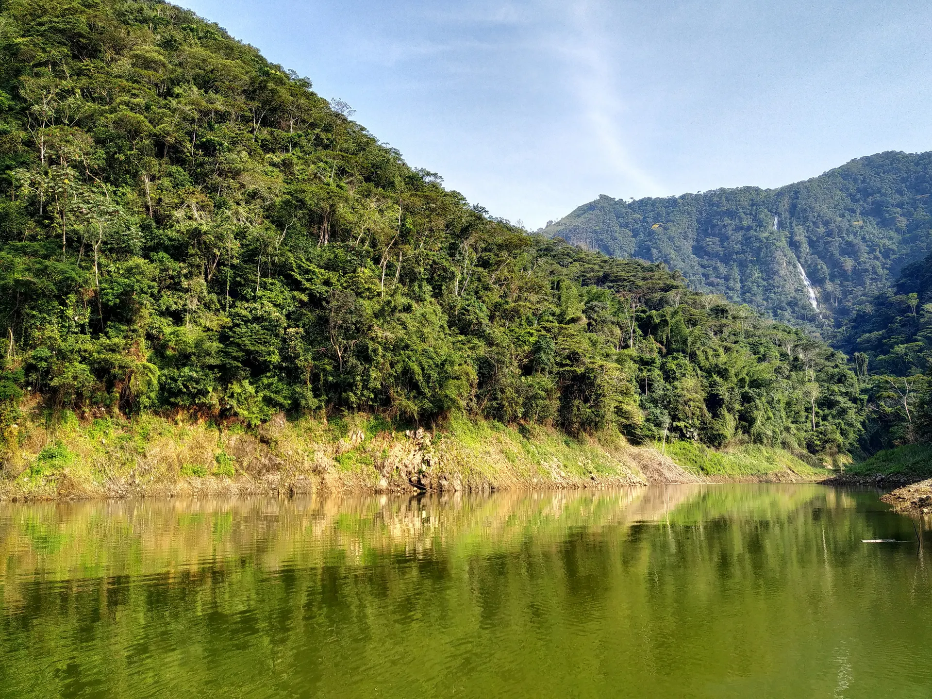 Laderas de selva muy tupida cayendo sobre un embalse en calma, con una cascada lejana asomando entre las montañas.