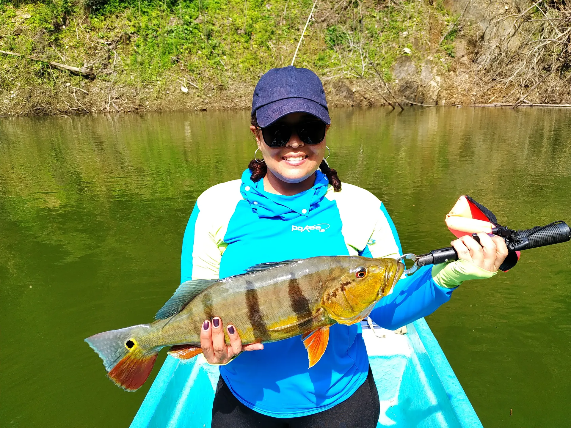 Una mujer con gorra y gafas de sol sostiene con las dos manos un pavón grande de rayas oscuras, en un bote sobre el embalse.