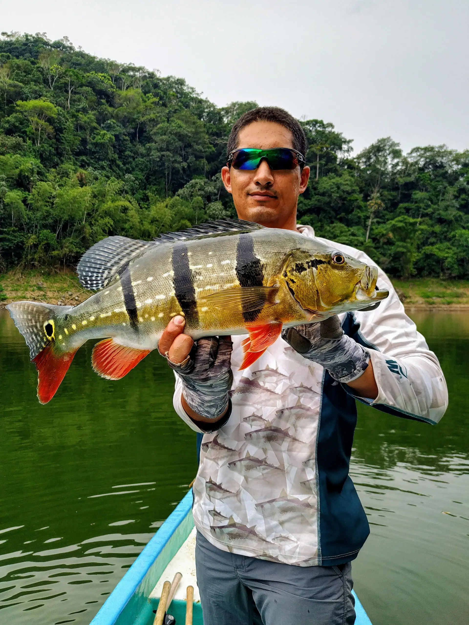 Un pescador con gafas de espejo sostiene con las dos manos un pavón grande de rayas negras y cola roja, en un bote sobre el embalse.