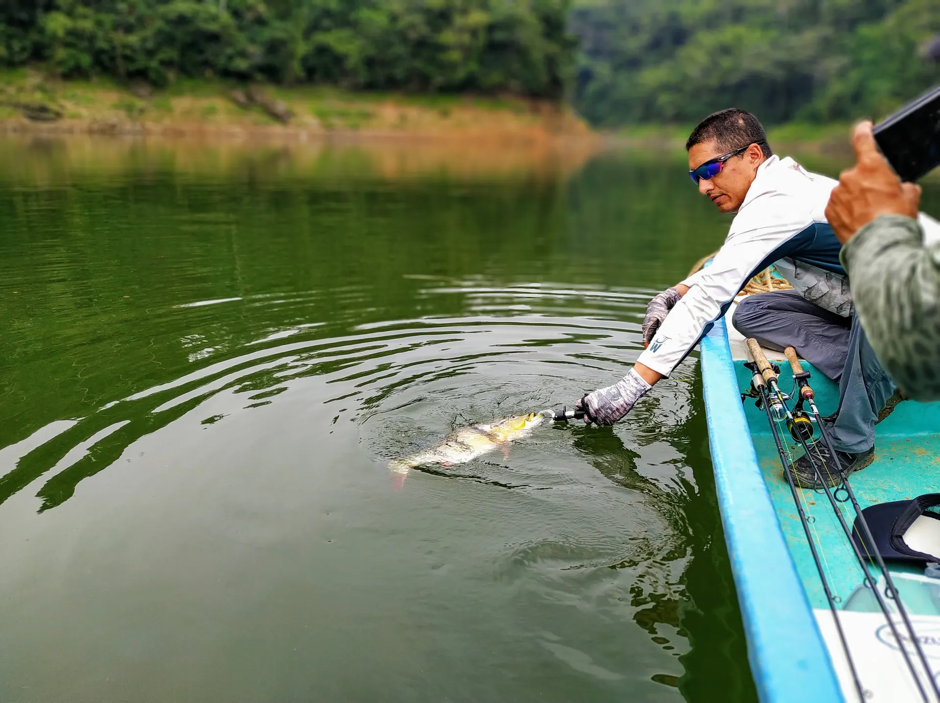 Un pescador se asoma por el borde del bote para devolver un pavón al agua, mientras otro compañero levanta el pulgar detrás.