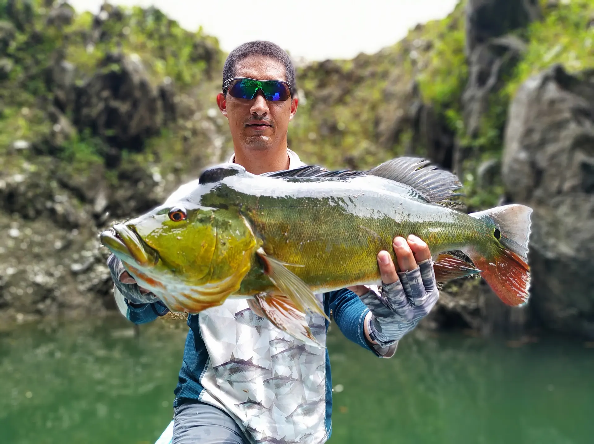 Un pescador sostiene con las dos manos un pavón grande de colores verde y dorado con la cola roja, frente a un paredón de roca cubierto de vegetación.