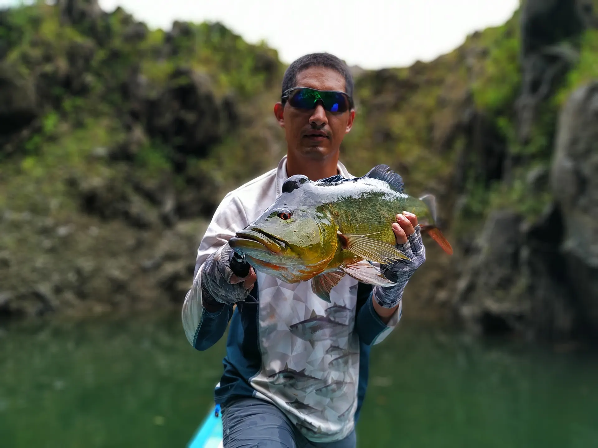 Un pescador con gafas de espejo sostiene un pavón dorado con las dos manos, frente a un paredón de roca cubierto de musgo.
