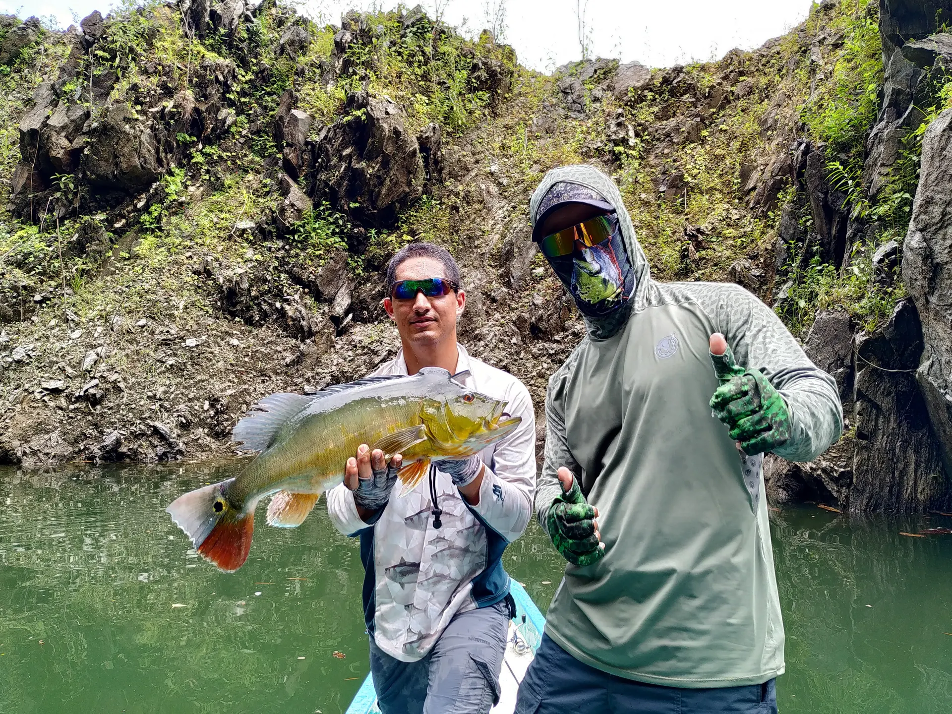 Dos pescadores posan junto a un pavón dorado: uno lo sostiene y el otro, con pasamontañas y gafas de espejo, levanta el pulgar.