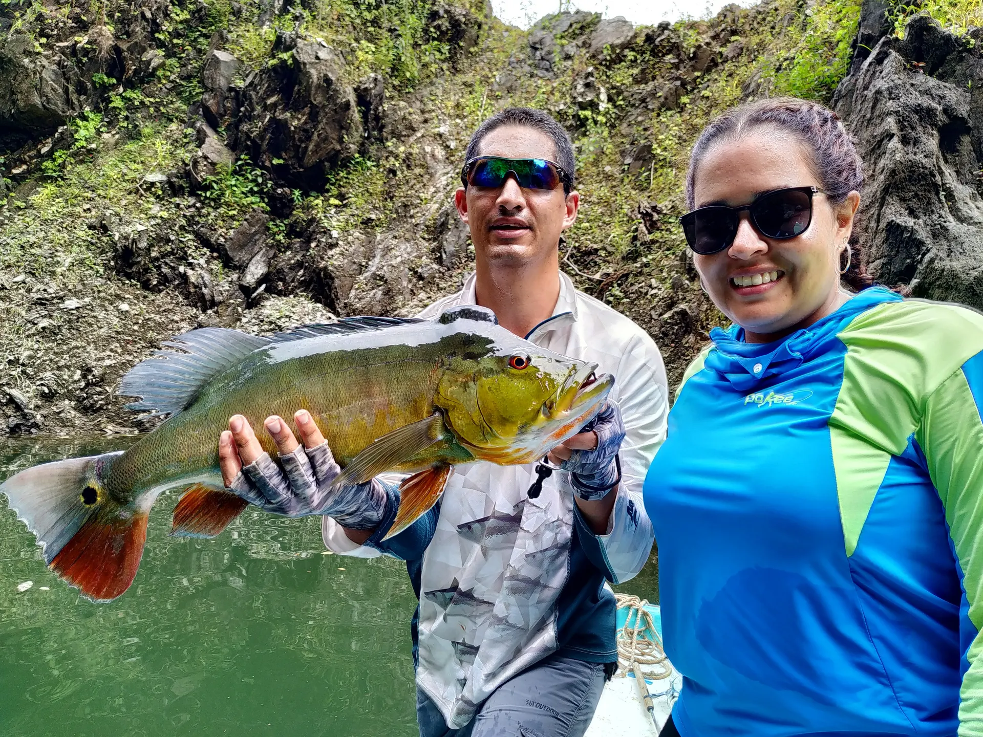 Un pescador sostiene un pavón dorado grande mientras una mujer con gafas de sol sonríe a su lado, frente a un paredón de roca.