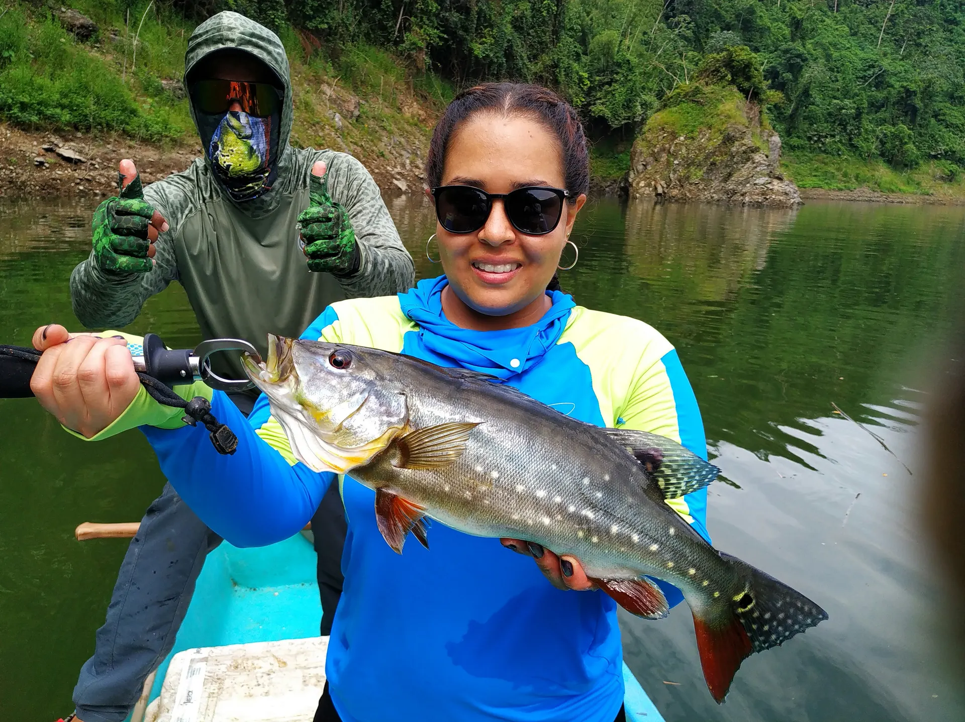 Una mujer sostiene un pavón grande de escamas plateadas mientras un compañero con pasamontañas levanta los pulgares detrás, en un bote sobre el embalse.