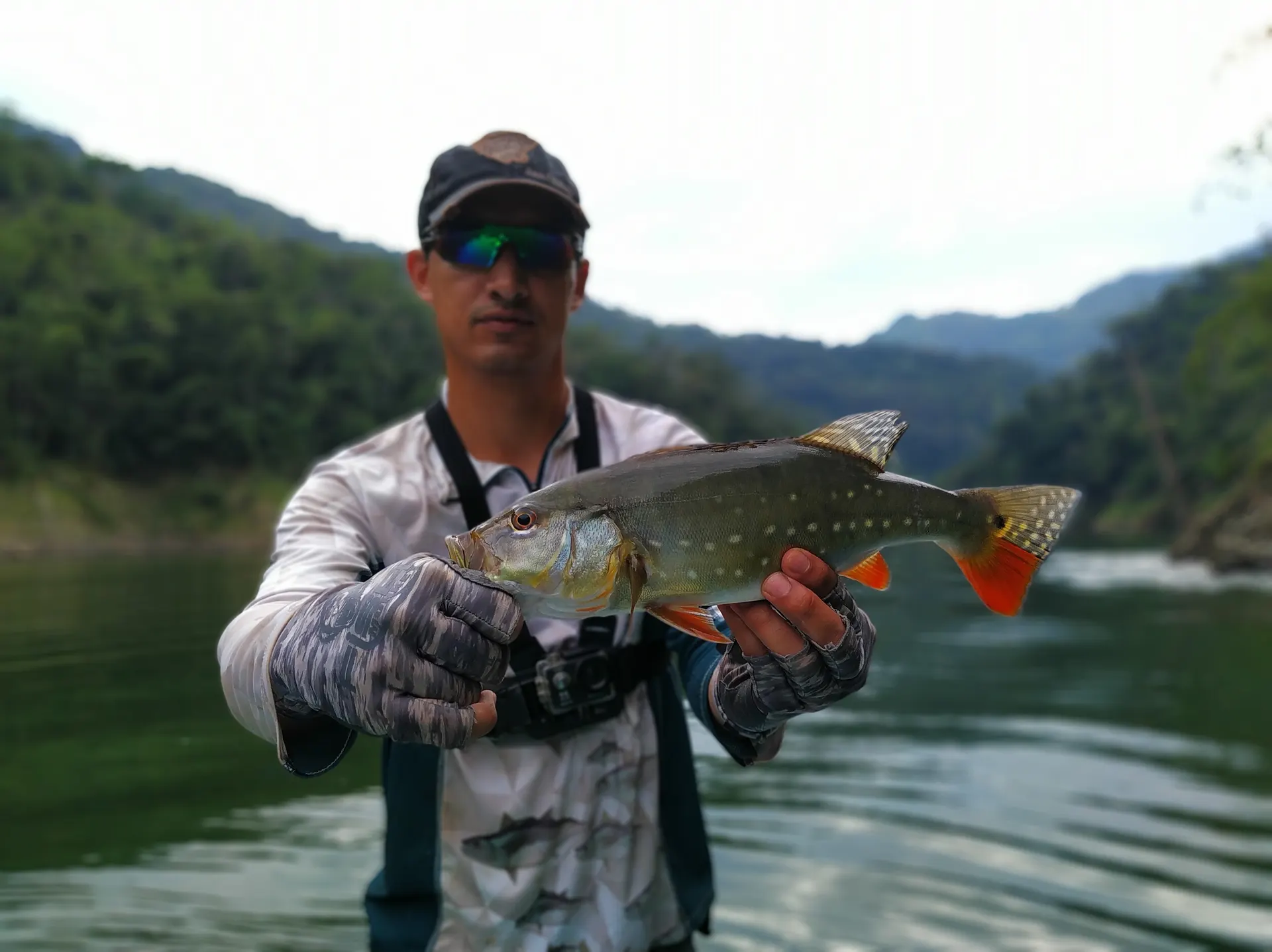 Un pescador con gorra y gafas de espejo sostiene un pavón de aletas anaranjadas, con montañas selváticas desenfocadas al fondo.